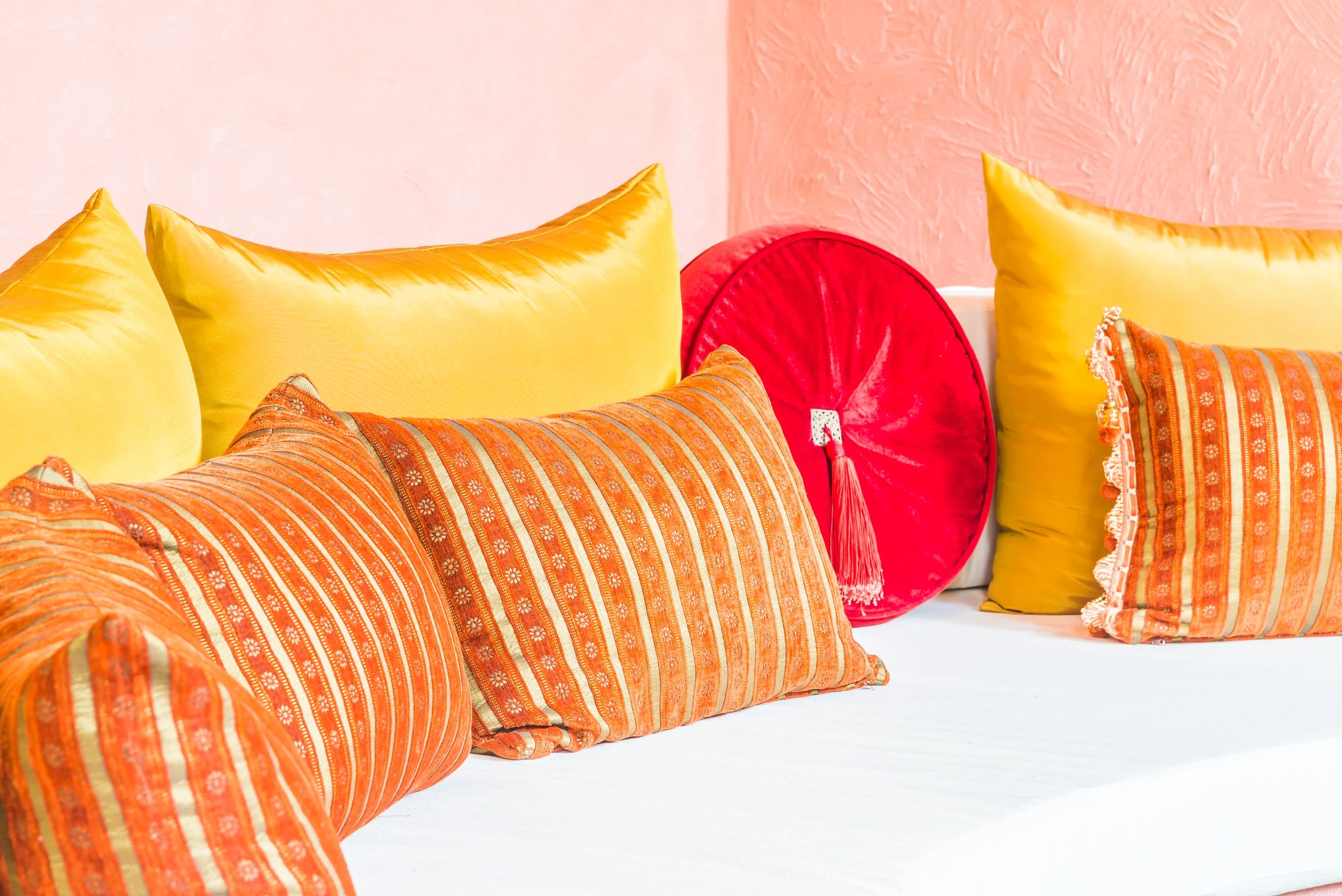 A row of colorful decorative pillows, including yellow, orange, and a red pillow with a tassel, on a white surface against a pink wall.