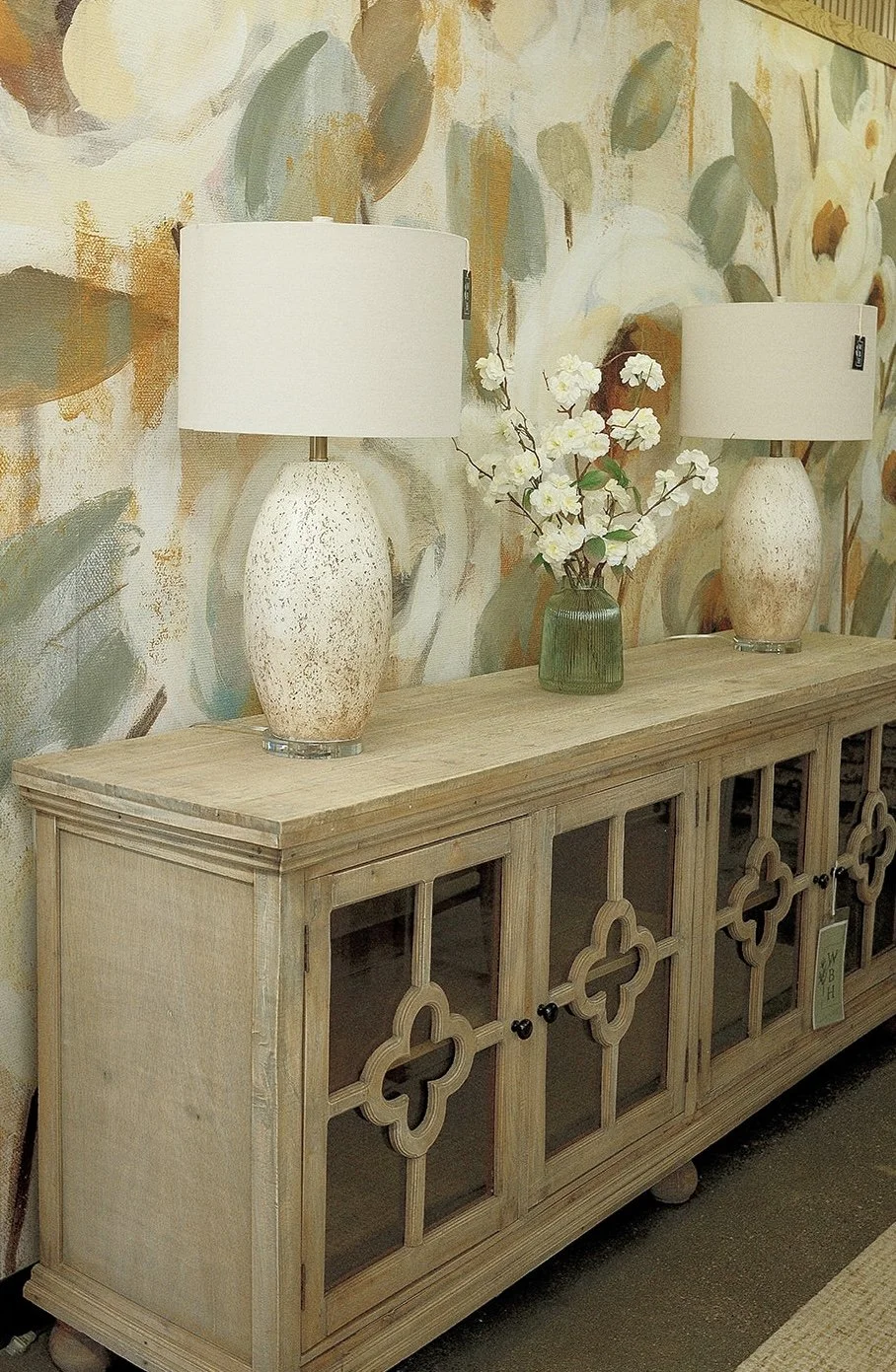 A wooden sideboard with decorative glass doors, topped with two large white lamps with textured bases and a green vase with white flowers, against a colorful abstract floral wallpaper background.