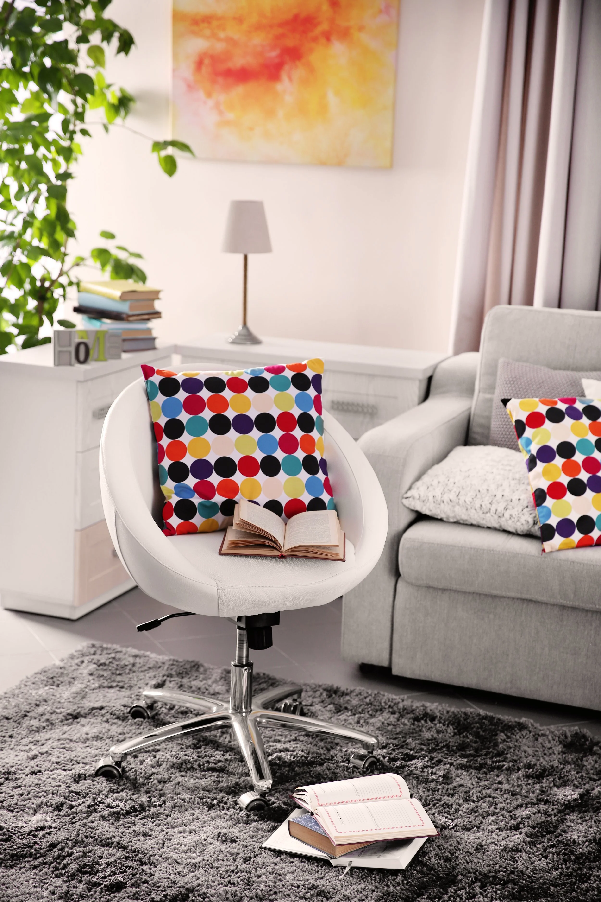 A modern living room with a gray sofa decorated with colorful polka dot pillows, an office chair with more polka dot pillows, an open book on the chair and on the gray rug, a white side table with stack of books and decorative blocks, a white dresser, a small lamp, a large colorful abstract painting, and a green plant.
