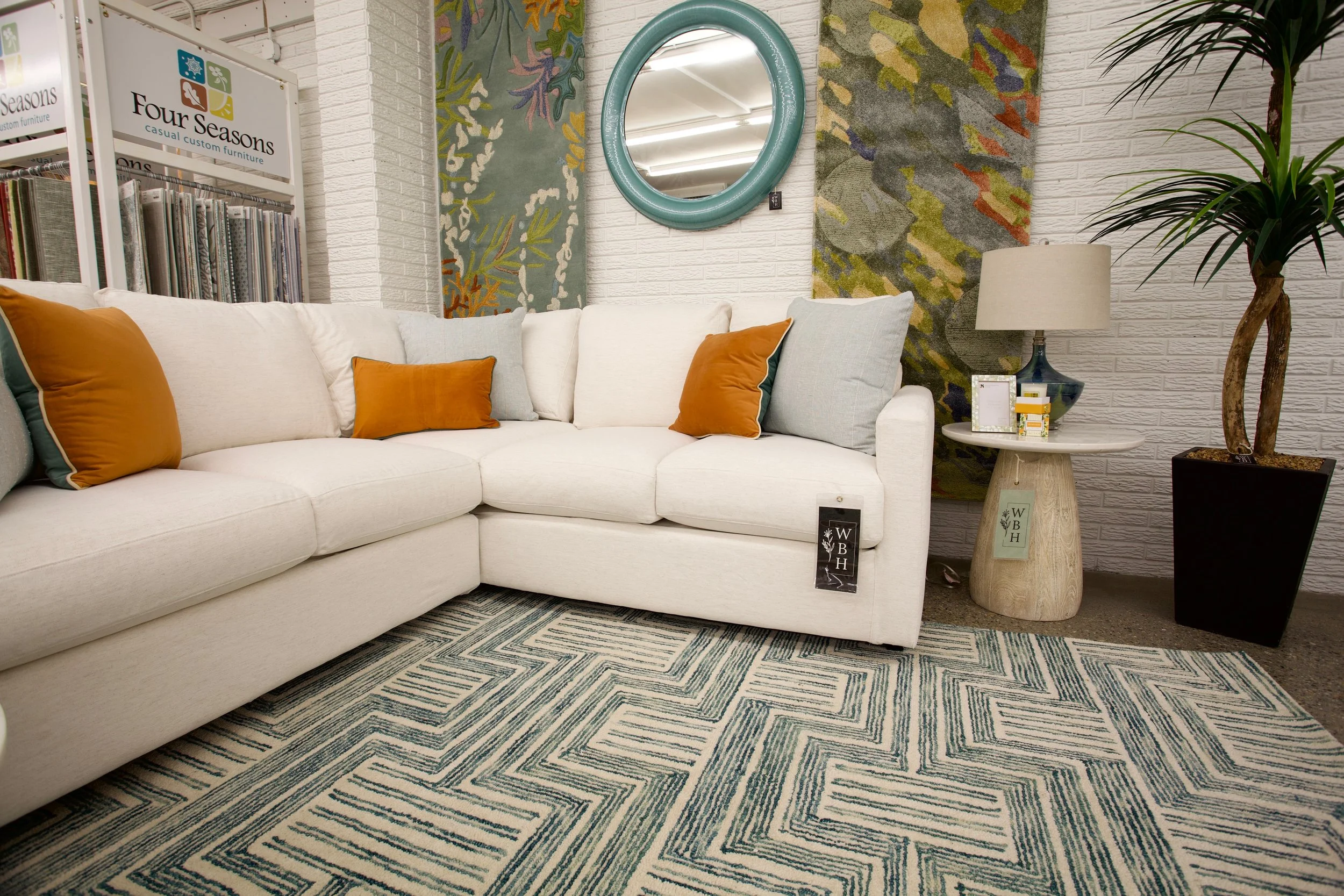 A white sectional sofa with orange and light green pillows in a furniture store. Behind the sofa, there are patterned rugs on the wall, a mirror, a table with a lamp, a potted plant, and a stack of fabric samples. There is a sign that reads "Four Sea