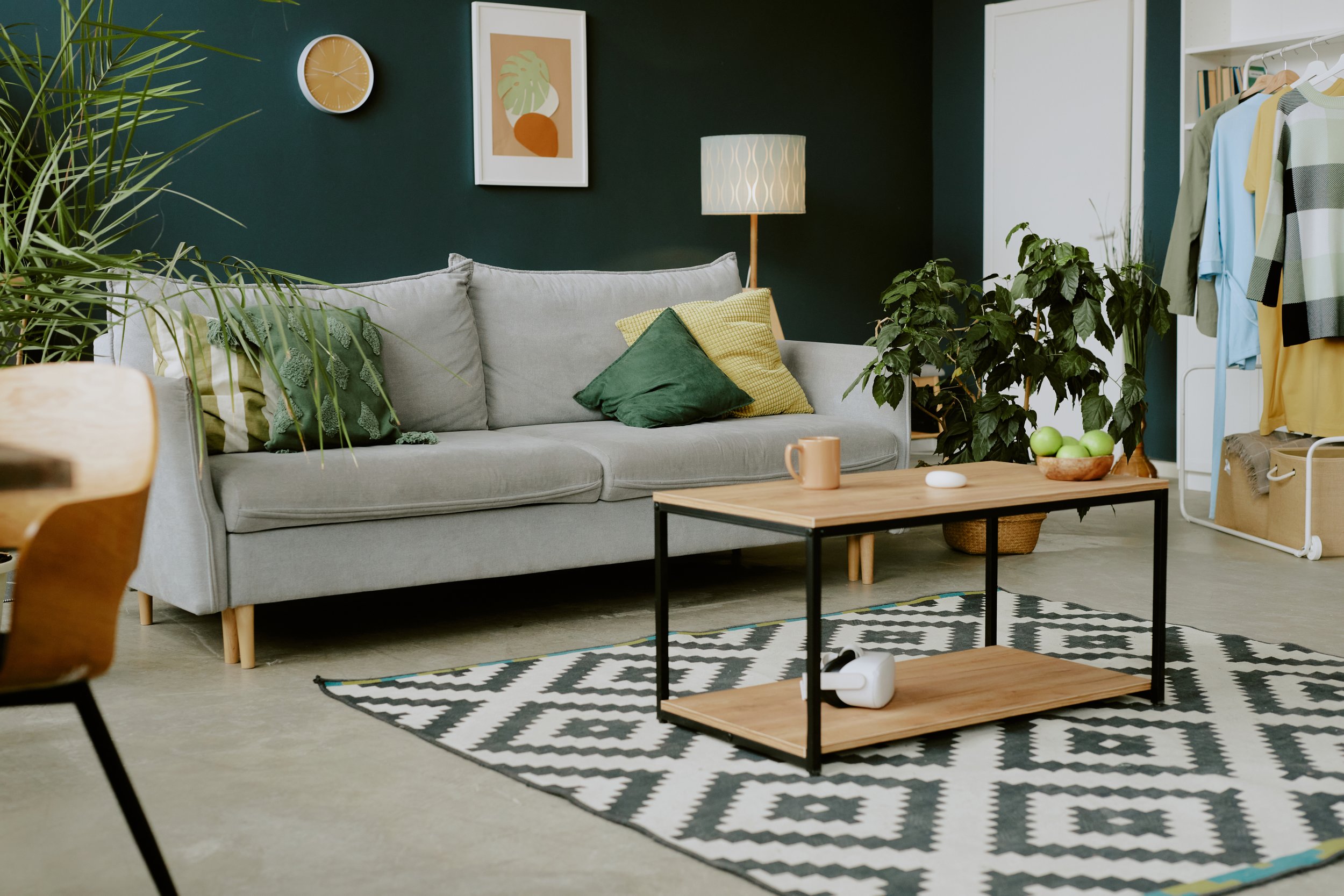 Living room with a light gray sofa, green and yellow pillows, a wooden coffee table with black metal frame, a patterned black and white rug, green plants, a floor lamp with a white lampshade, wall art, a round wall clock, and a white clothing rack with hanging clothes