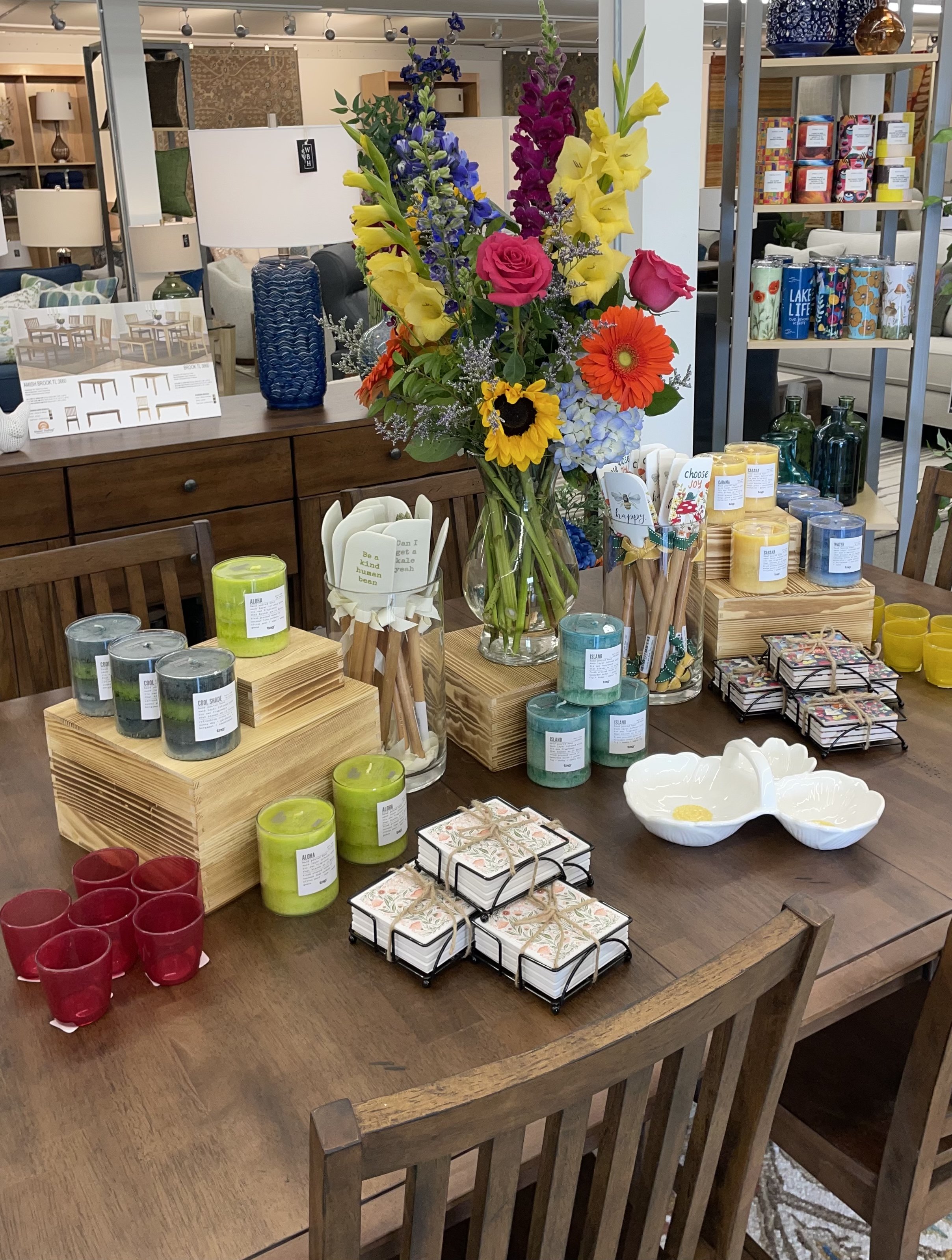 A retail store display featuring a bouquet of colorful flowers in a glass vase, surrounded by candles, decorative items, and small gift boxes on a wooden table.