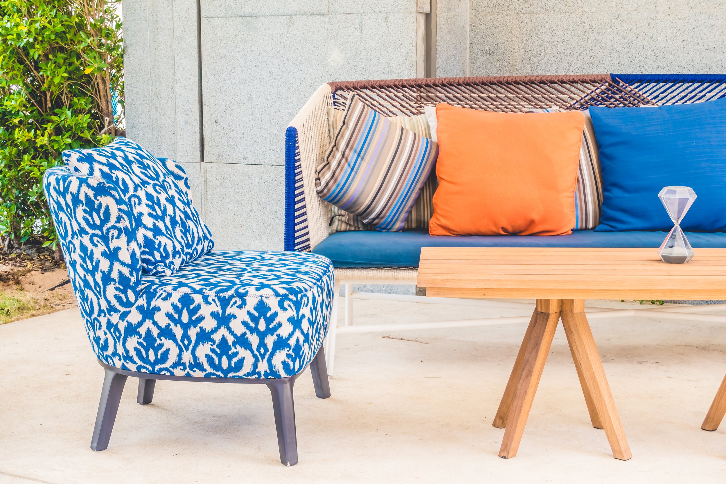 Outdoor patio seating with a blue patterned chair, a wooden table, and a wicker sofa with colorful pillows, including orange, blue, and striped designs, and a decorative hourglass on the table.
