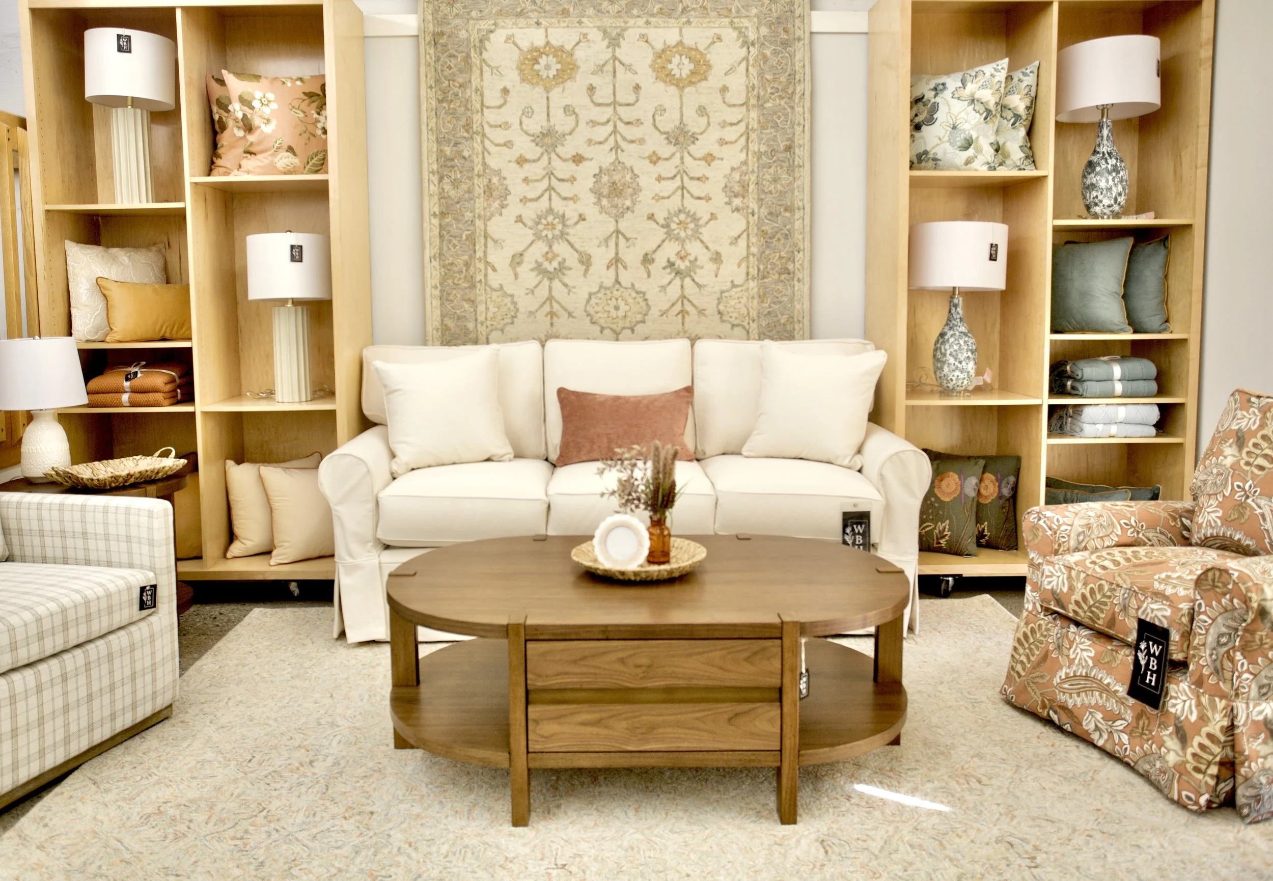 A living room with a white sofa, a wooden coffee table, and shelves filled with pillows and lamps, with a patterned rug on the floor.