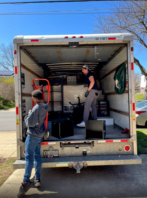 Two boys unloading or organizing items inside the back of a moving truck on a sunny day.