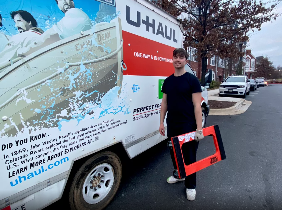 A young man standing next to a U-Haul truck on a street, holding a red-framed pocket saw, wearing a black t-shirt, black pants, and white sneakers, with parked cars and residential buildings in the background.