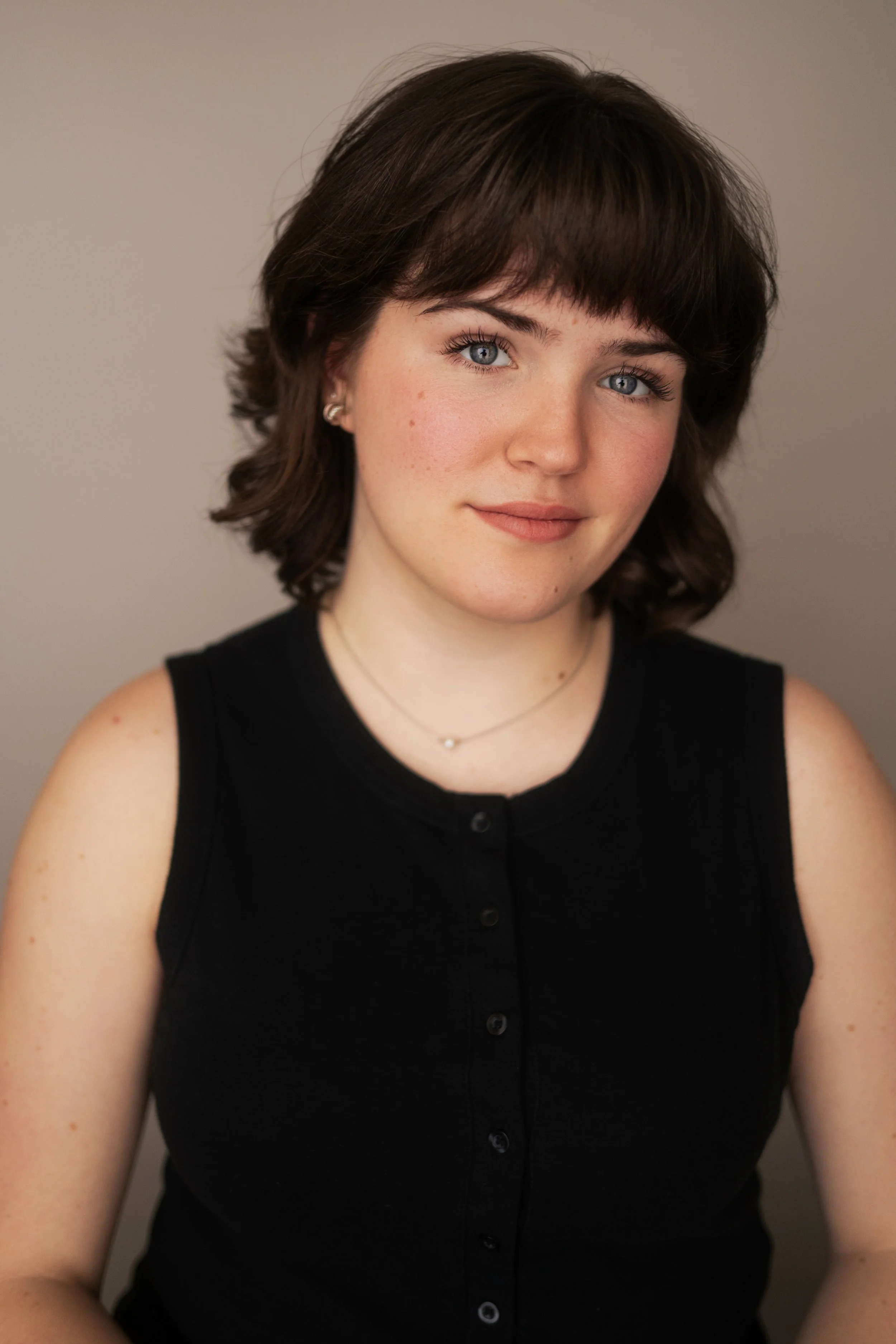A young woman with short dark brown hair and blue eyes, wearing a sleeveless black top, standing against a plain light-colored background.