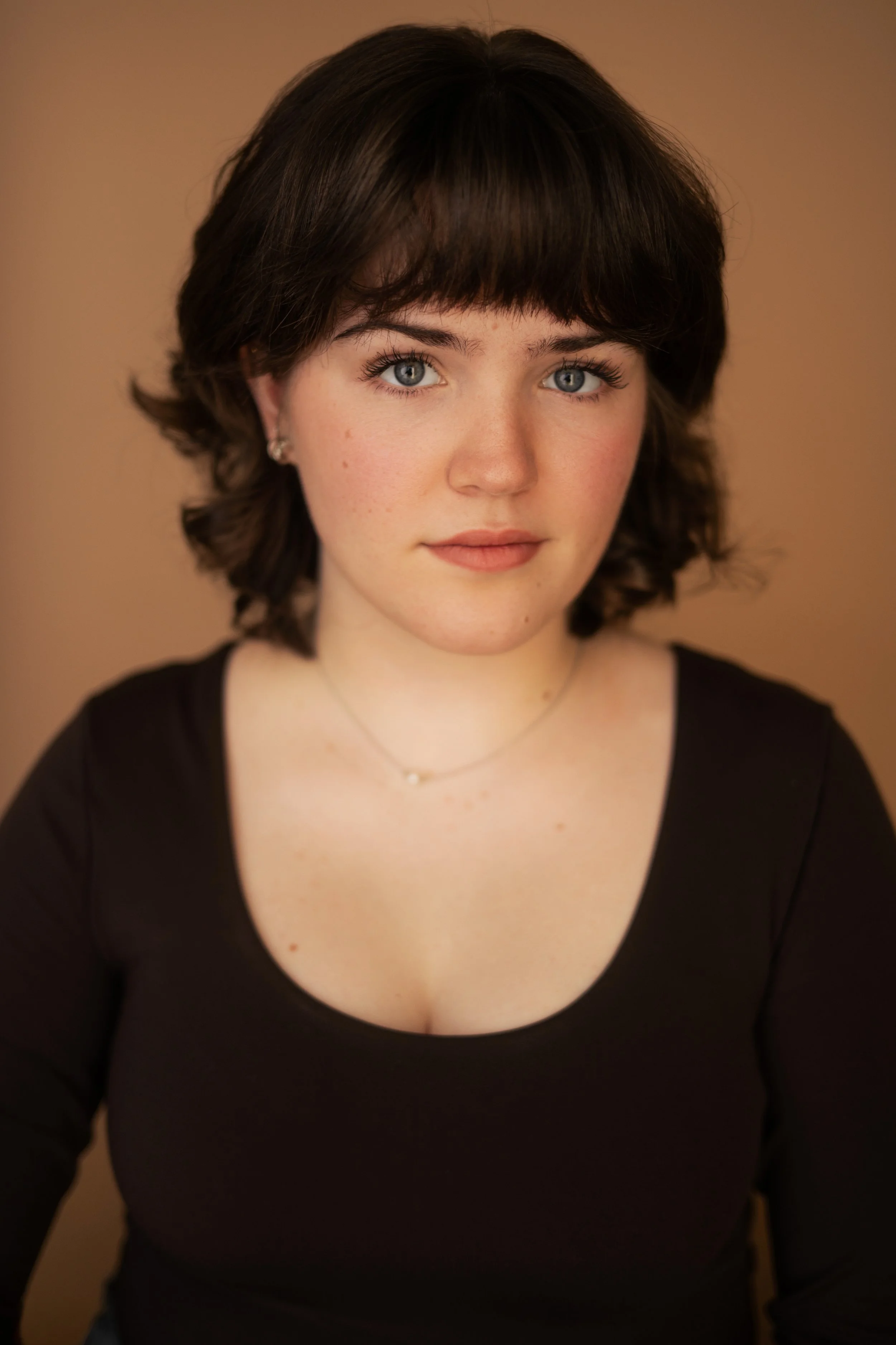 Close-up portrait of a young woman with short brown hair, blue eyes, and fair skin wearing a black top and a delicate necklace, against a warm, neutral background.