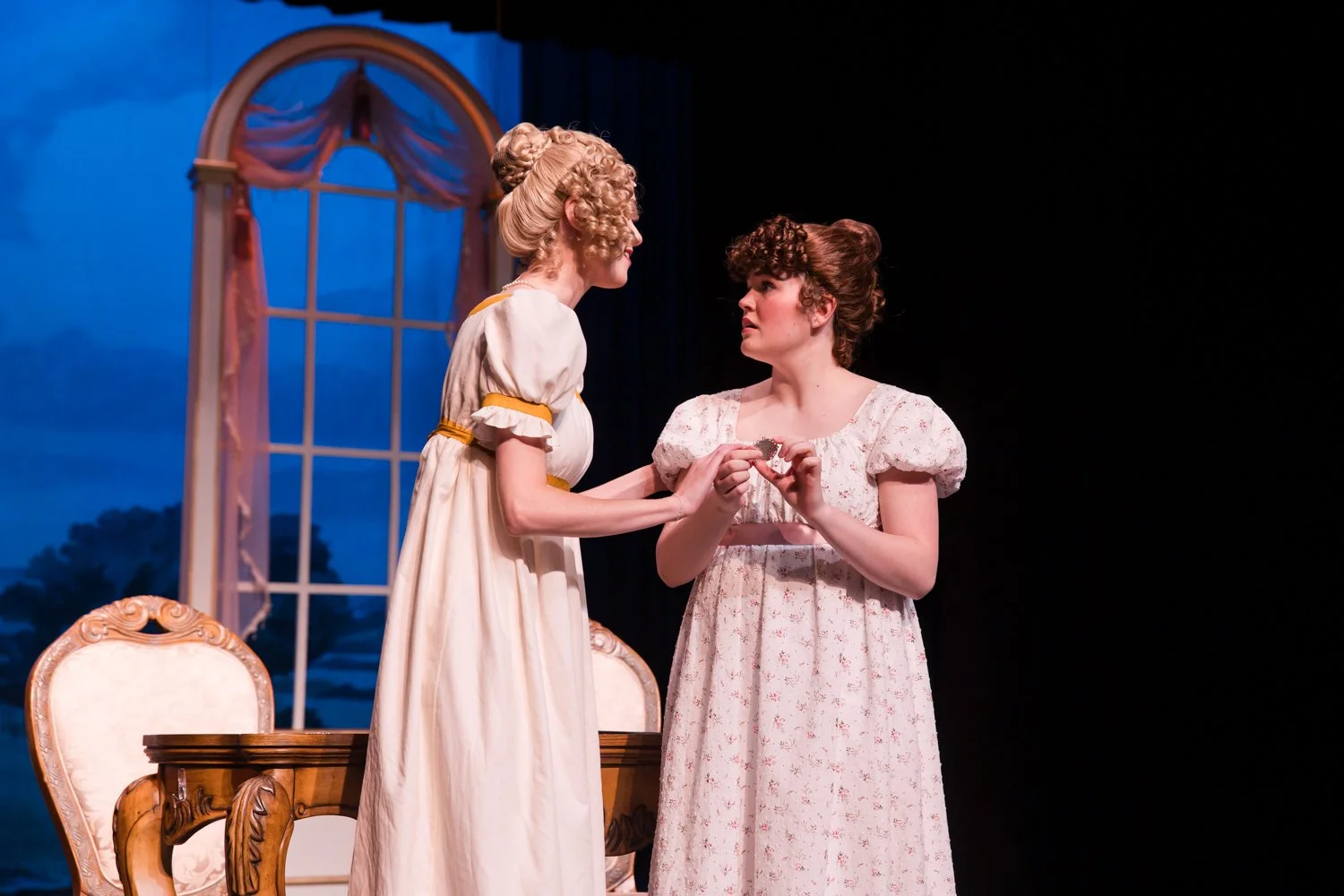 Two women in period dresses on stage, one standing and leaning forward, the other standing with hands clasped, during a theatrical performance with a painted window backdrop.
