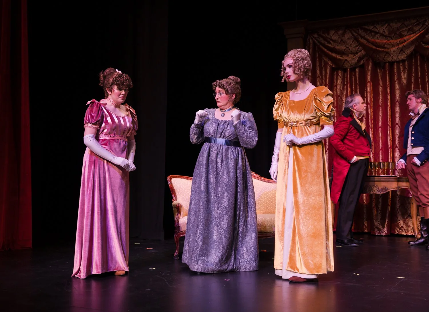 Actors on stage dressed in vintage costumes, with three women in the foreground and two men in the background, in a theatrical play setting.