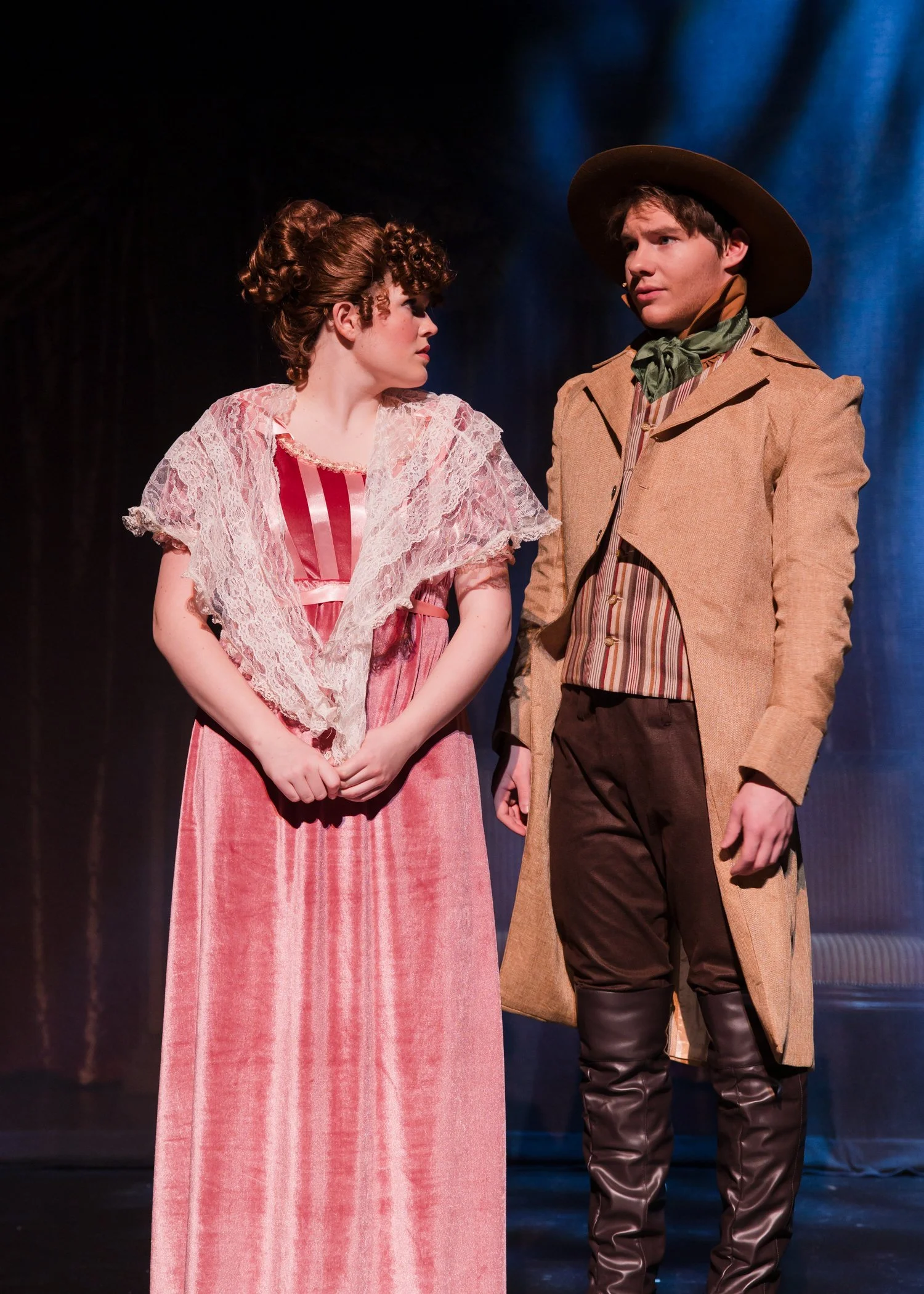 Two actors on stage in period costumes, a woman in a pink dress with lace shawl and a man in cowboy attire, engaged in a theatrical scene.