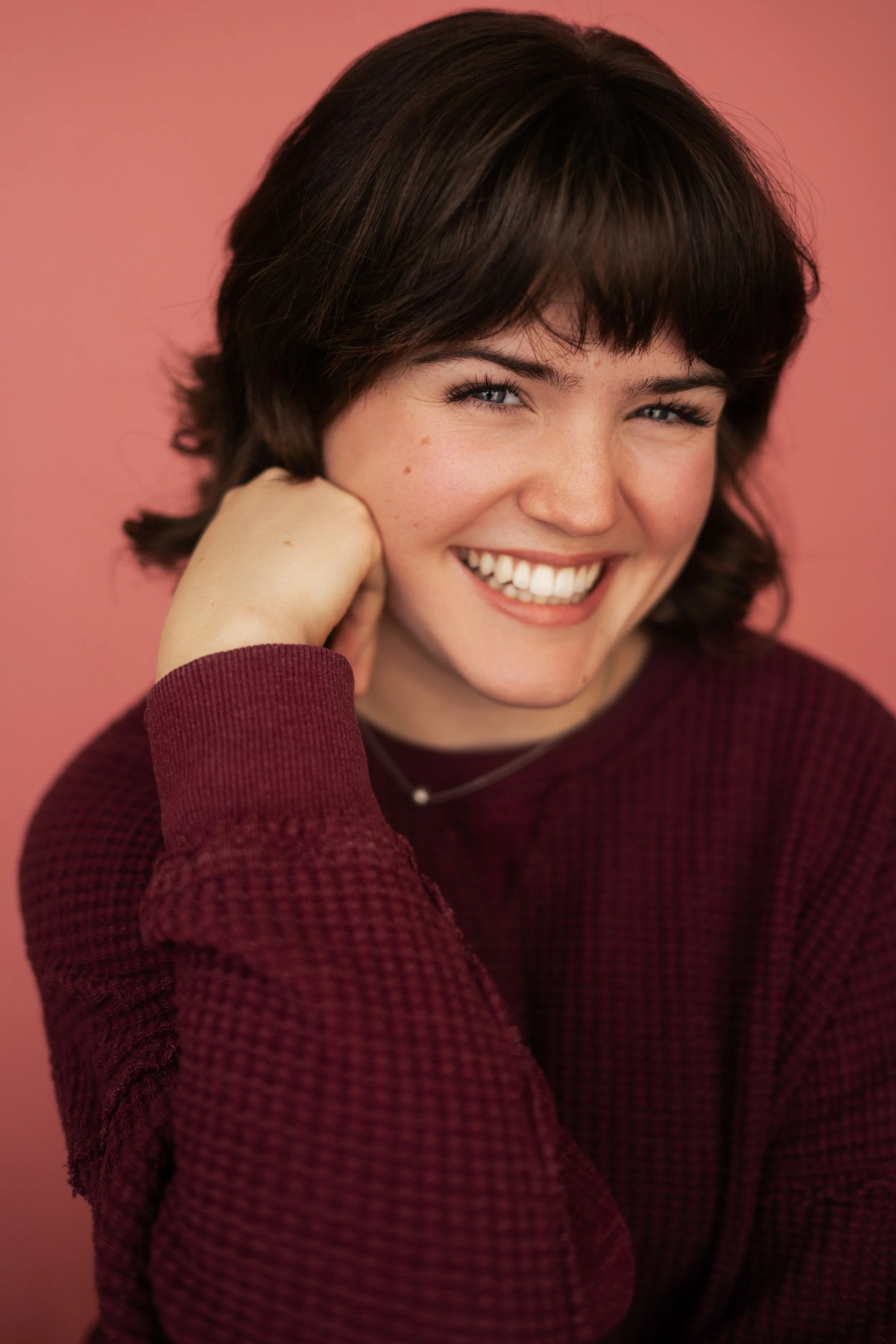Young woman with short brown hair, smiling, wearing a maroon sweater, against a pink background.