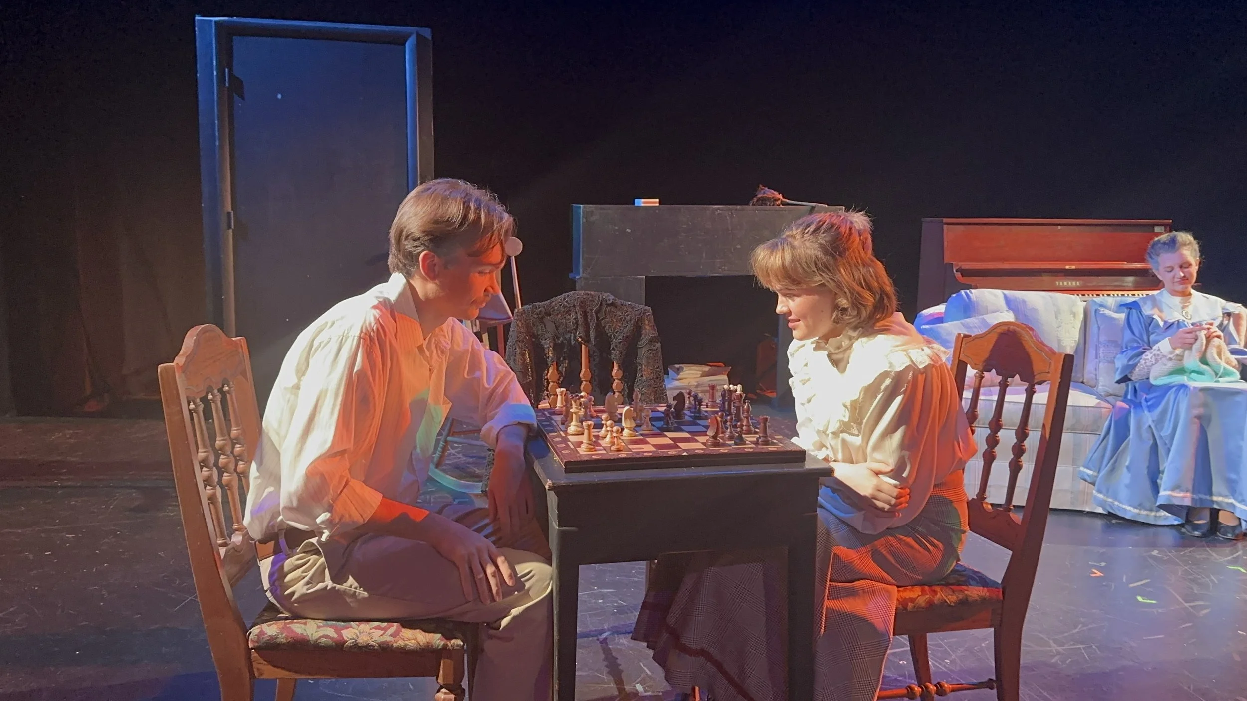 Two people playing chess at a wooden table with two other women seated in the background, all dressed in vintage clothing, on a stage set resembling a living room.