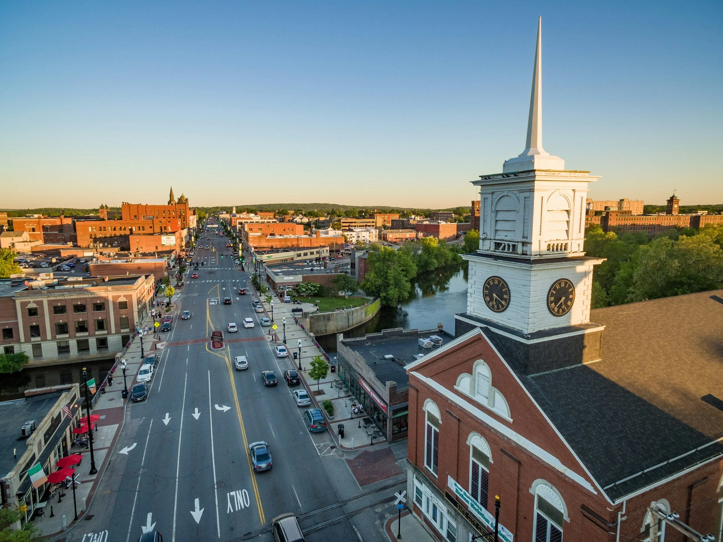 Aerial view of a small downtown with a prominent white clock tower on a red brick building, streets with cars, trees, and a river running through the city at sunset.