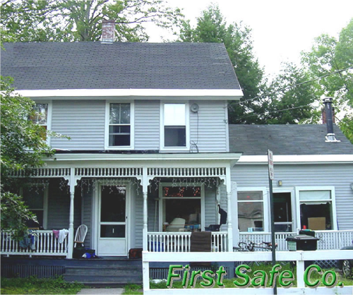 Front view of a light blue two-story house with a porch, surrounded by trees, with the words "First Safe Co." in green at the bottom right.