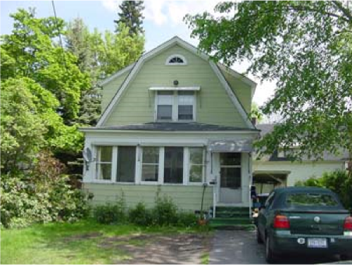 A light green, two-story house with a front porch, surrounded by greenery and trees, and a black car parked in the driveway.