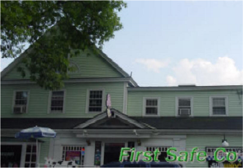A two-story green building with a gabled roof and multiple windows, partially obscured by a tree, with a sign reading 'First Safe Co.'