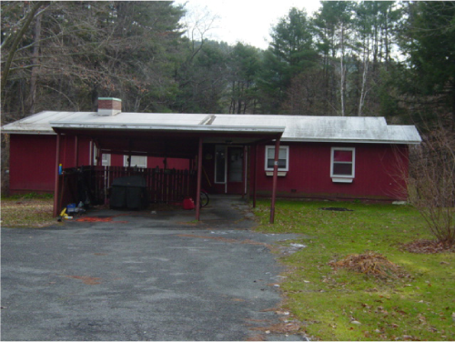Single-story red house with white roof, located in a rural area surrounded by trees, with a small driveway and a covered porch.