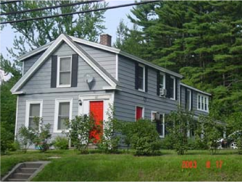 A two-story house painted gray with black shutters and a bright red front door, surrounded by green bushes and trees.