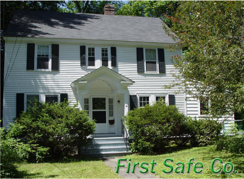A two-story white house with black shutters and a gabled porch, surrounded by green bushes and trees, with the text 'First Safe Co.' in green at the bottom right corner.