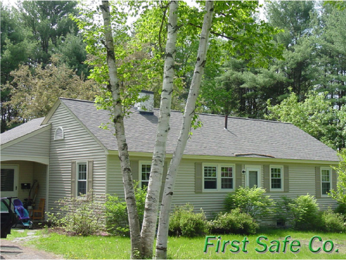 A gray house with a sloped roof and multiple windows surrounded by trees and greenery, with the words "First Safe Co" in the lower right corner.