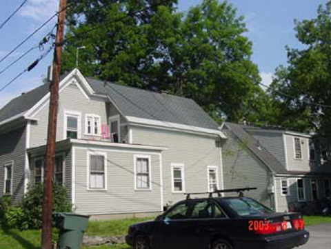 A two-story white house with a gray roof, trees in the background, and a black car parked in front.