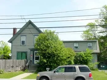 A two-story gray house with a gabled roof, front porch, and surrounded by trees and greenery. There is a silver SUV parked on the street in front of the house, and power lines overhead.