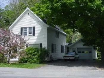 A white two-story house with a garage, surrounded by green trees and shrubs, with a car parked in the driveway.