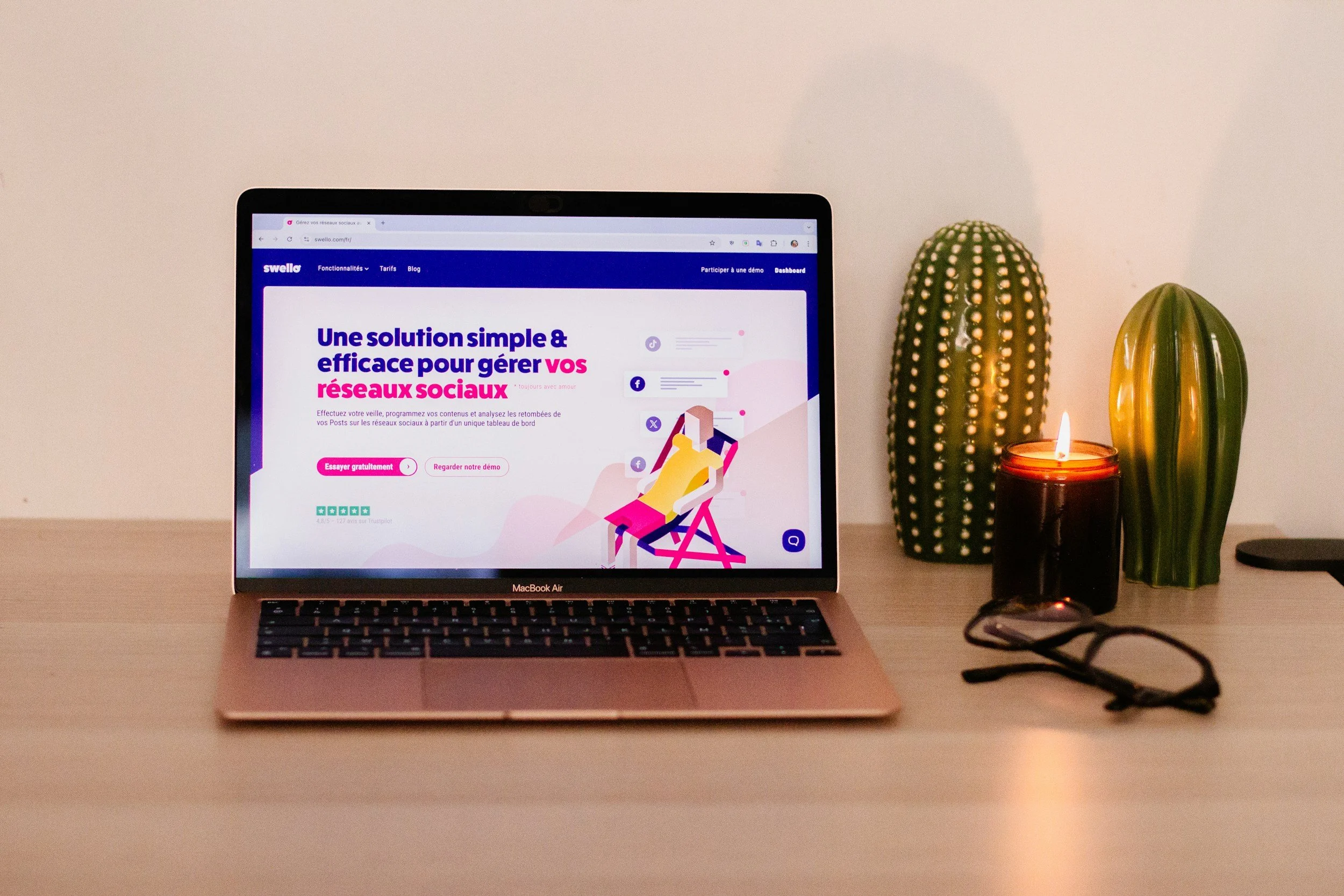 A MacBook Air on a desk displaying a website in French about social media management solutions, with two decorative cactus plants, a lit candle, and a pair of glasses on the desk.