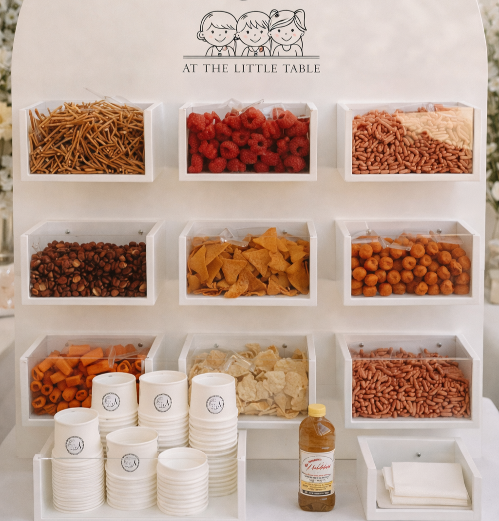 Snack station with various snacks in white containers, including pretzels, raspberries, pasta, nuts, tortilla chips, cheese puffs, carrot sticks, cauliflower, and pretzels. Cups and napkins are available for serving, and a bottle of iced tea is on the table.