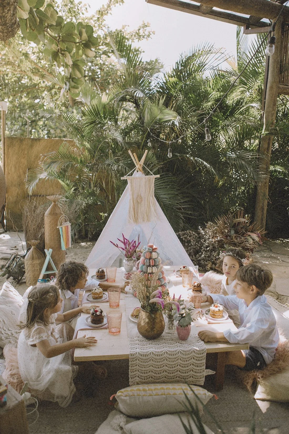 Children dressed in white celebrating a birthday or special occasion outdoors with decorated table, cakes, flowers, and a teepee in a lush garden setting.