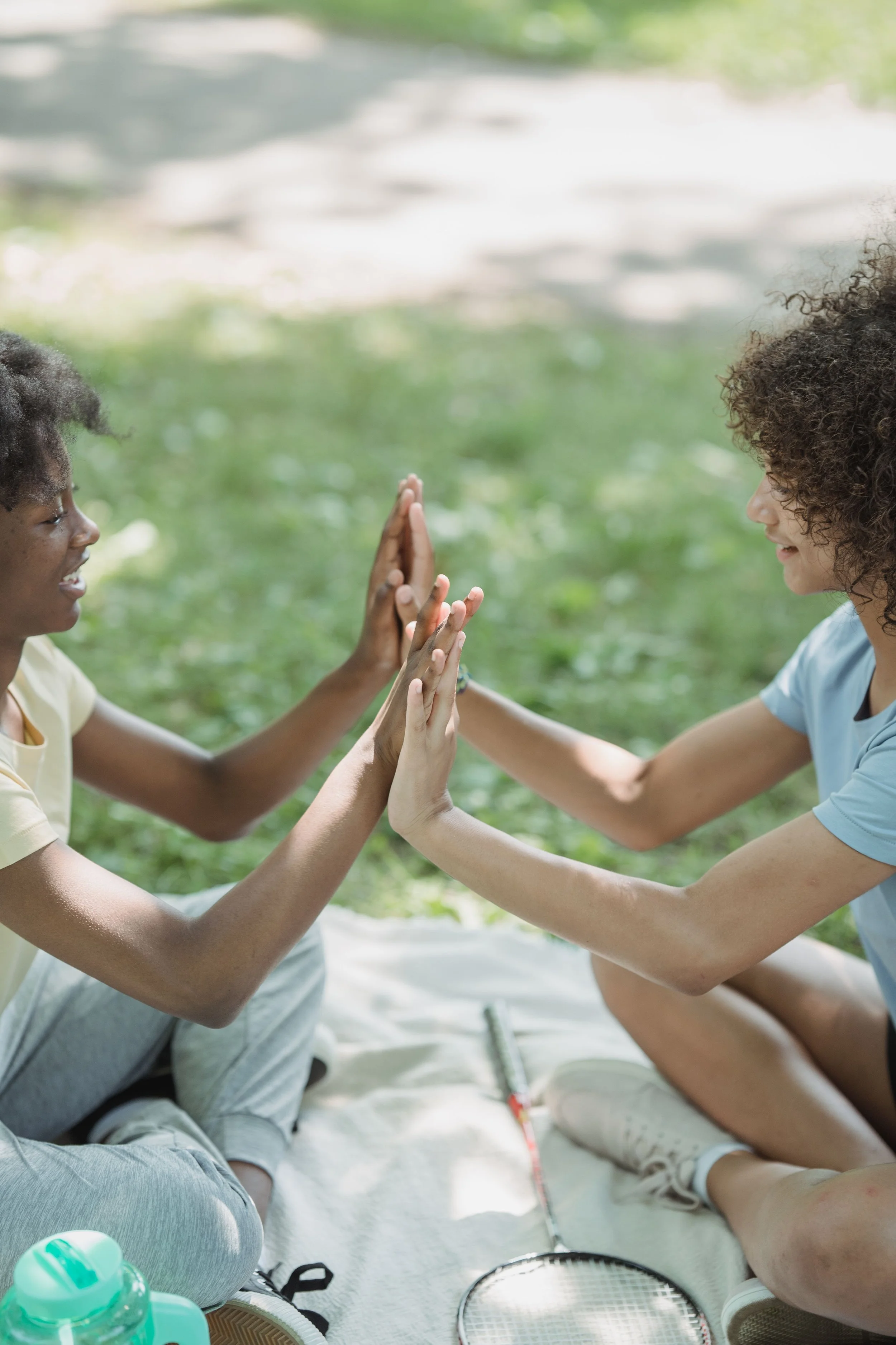 Two young women with curly hair sitting on the ground in a park, engaging in a high five. They are smiling and facing each other, wearing casual clothing, with tennis rackets and water bottles nearby.