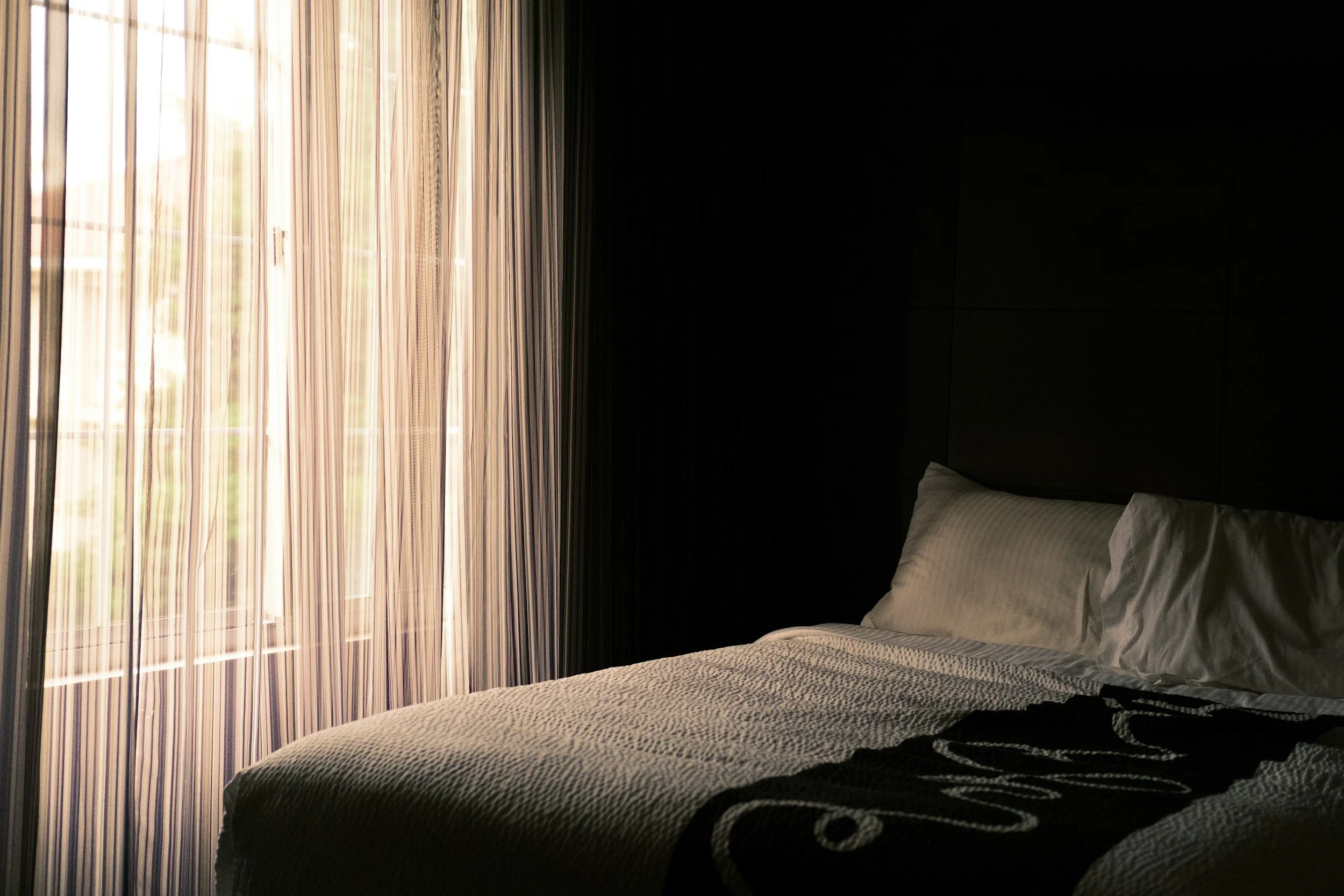 A neatly made bed in a dimly lit room with striped bedding and pillows, beside a window with beige curtains letting in natural light.