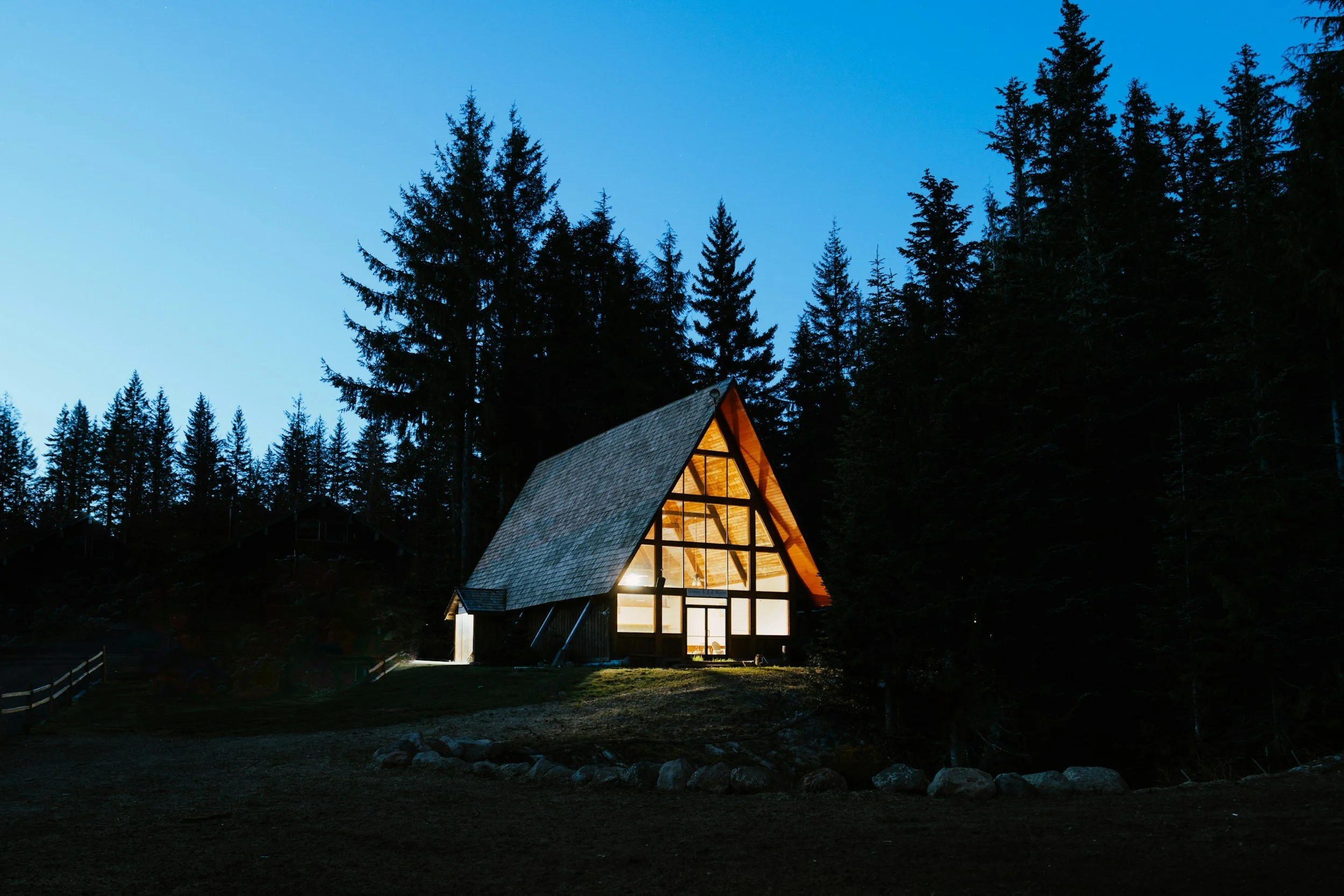 Aisle with classic ocean blue sky and trees and A-frame cabin with interior lights on, surrounded by nature in the evening