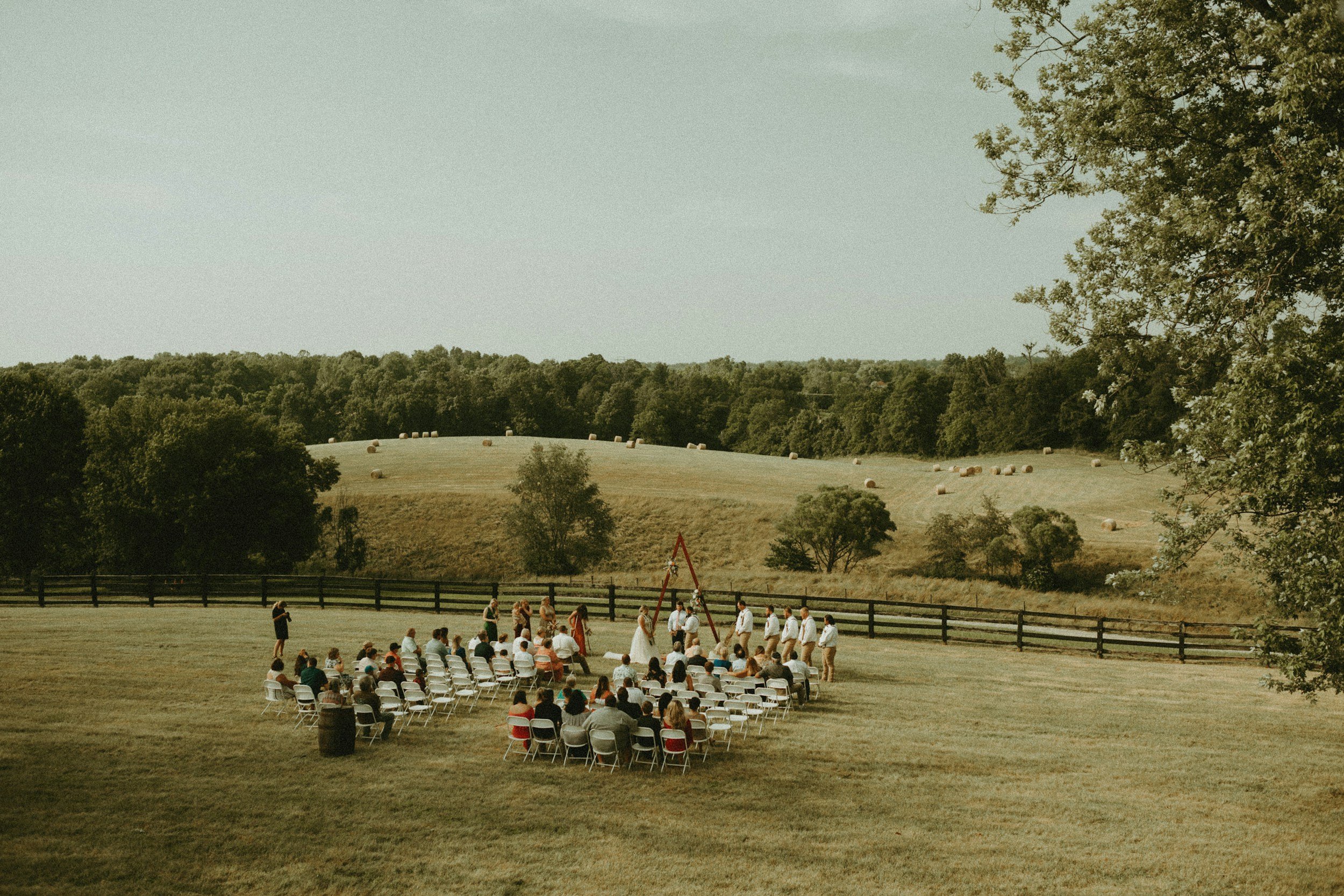 An outdoor wedding ceremony setup in a grassy area with chairs arranged in rows, a fence, and people gathered, with a hill and trees in the background.