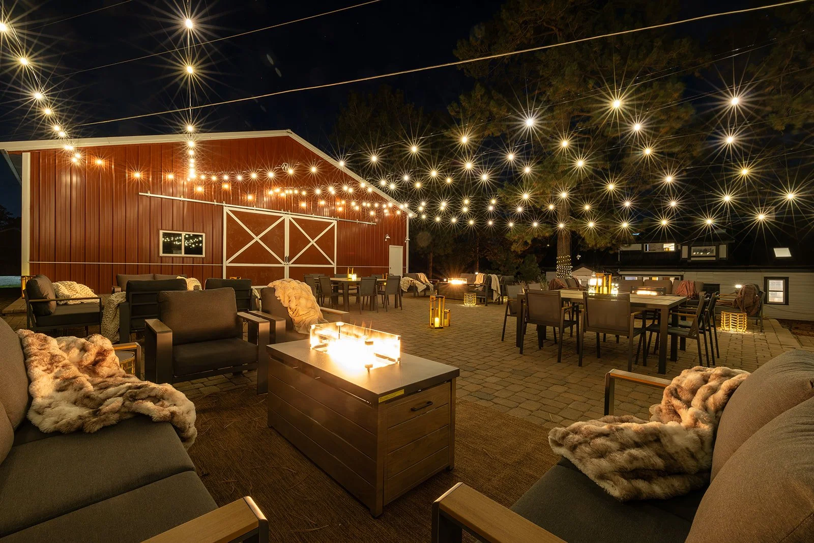 Nighttime outdoor patio decorated with string lights, featuring multiple seating areas with chairs, sofas, and throws, surrounded by trees and a red barn-style building.