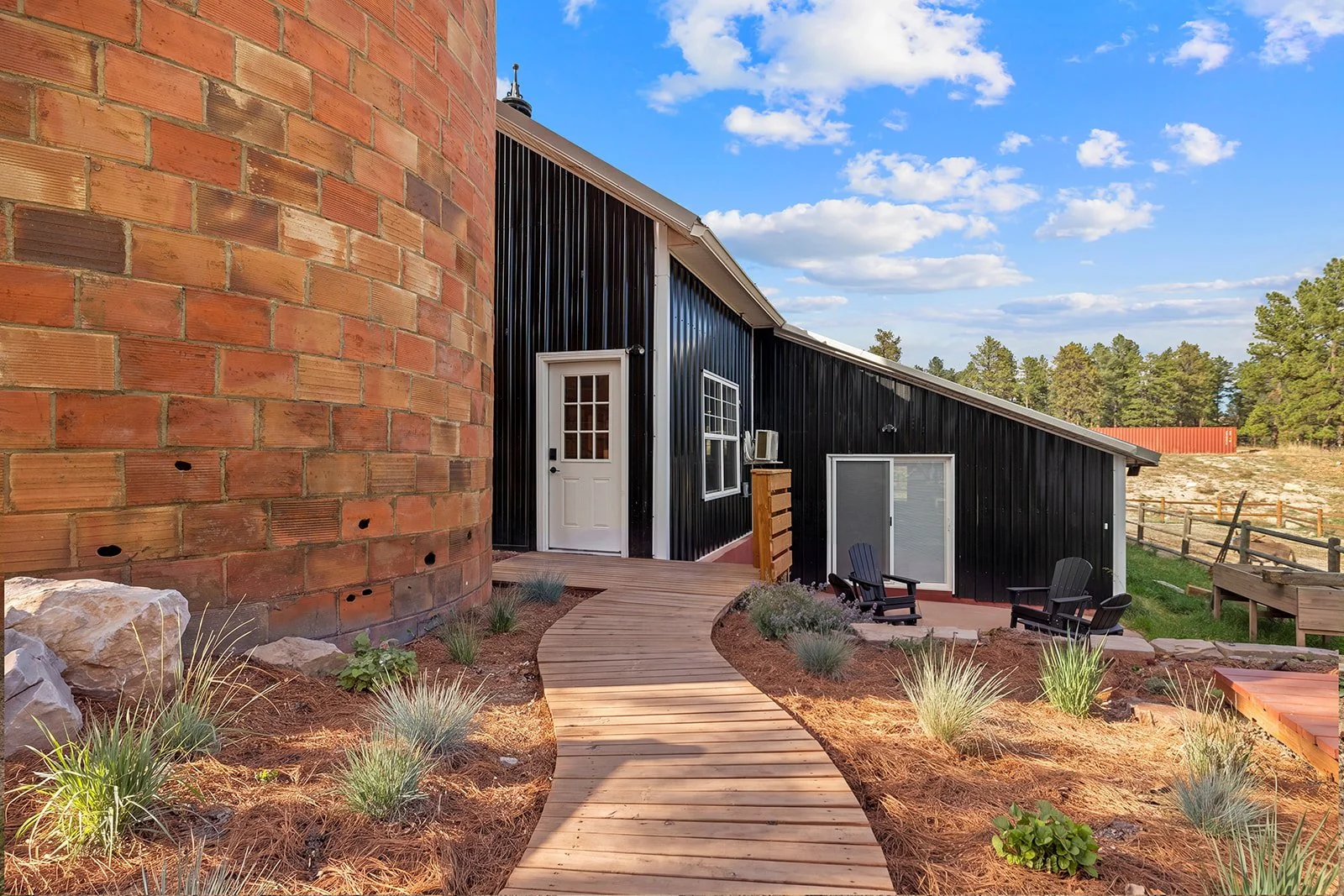 A modern house with black metal siding, a white door, and windows, with a landscaped yard featuring a wooden pathway, outdoor chairs, and a red brick chimney, under a partly cloudy sky.