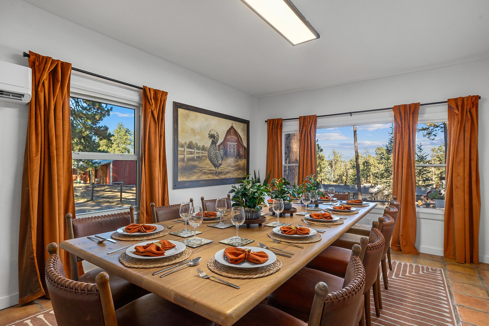 A dining room with a large wooden table set with plates, silverware, and orange napkins, surrounded by brown chairs, and decorated with two green potted plants in the center. The room has three large windows with orange curtains, a framed painting of a chicken and barn on the wall, and views of trees and a bright blue sky outside.