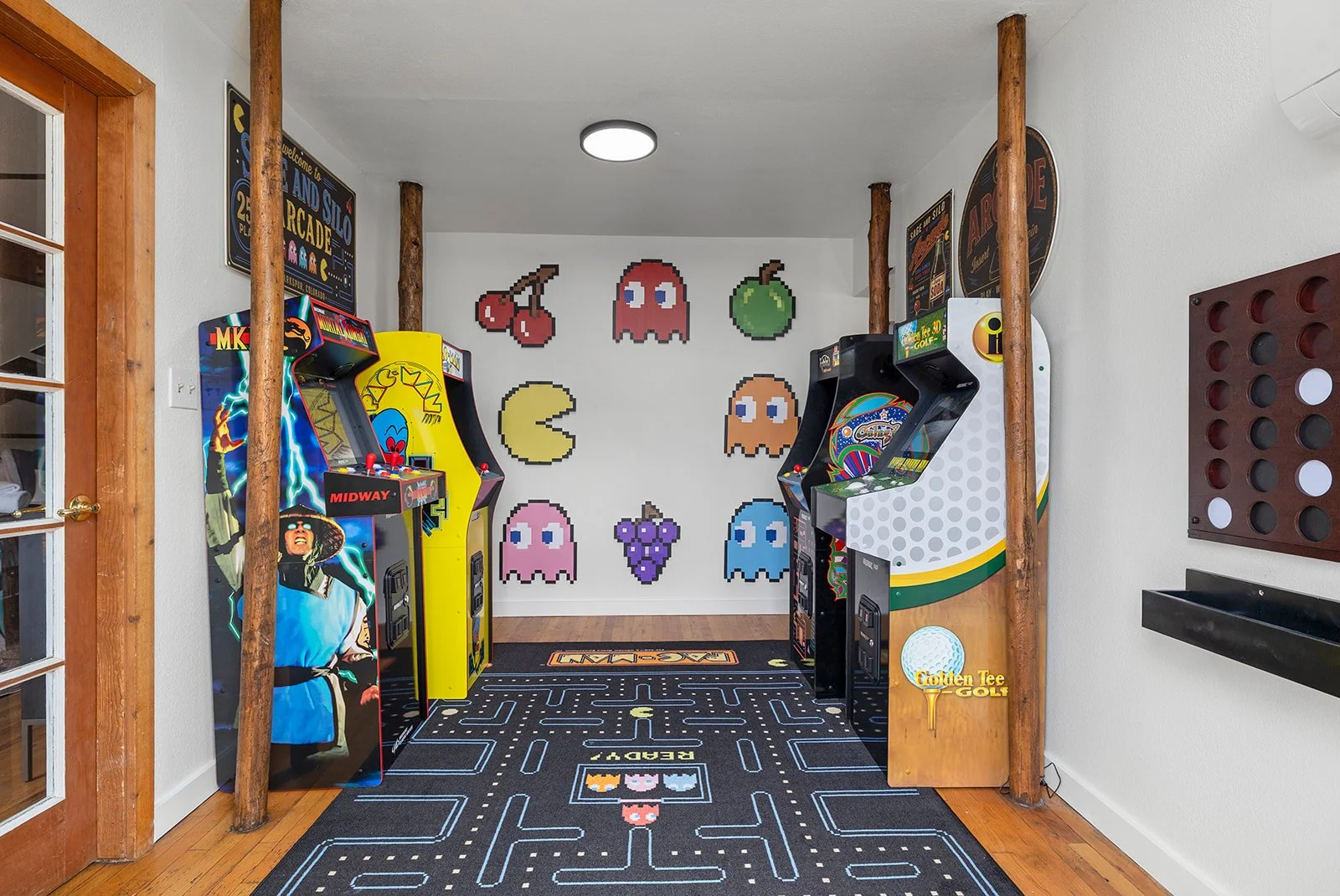 A vintage arcade gaming room with pixel art characters on the wall, two arcade game machines on either side, and a Pac-Man themed rug on the wooden floor.