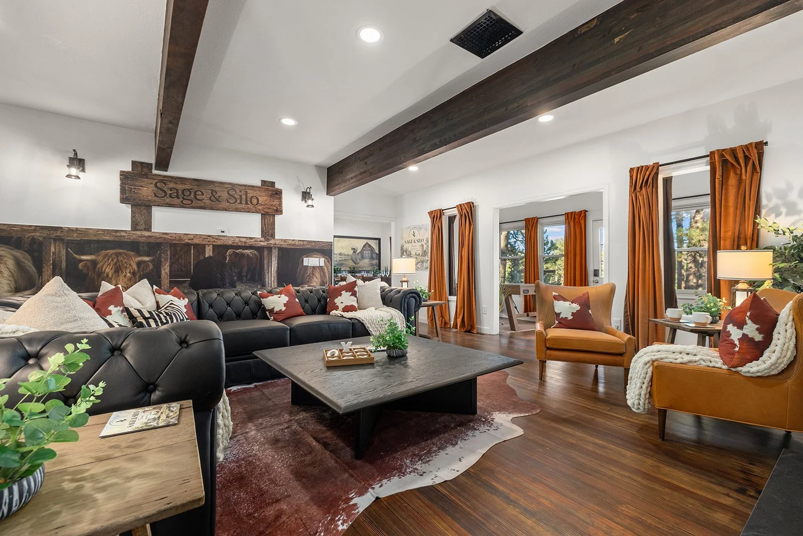 Living room with large black leather sofa, tan armchairs, orange curtains, wooden flooring, and rustic decor including a sign that says "Sage & Silo."