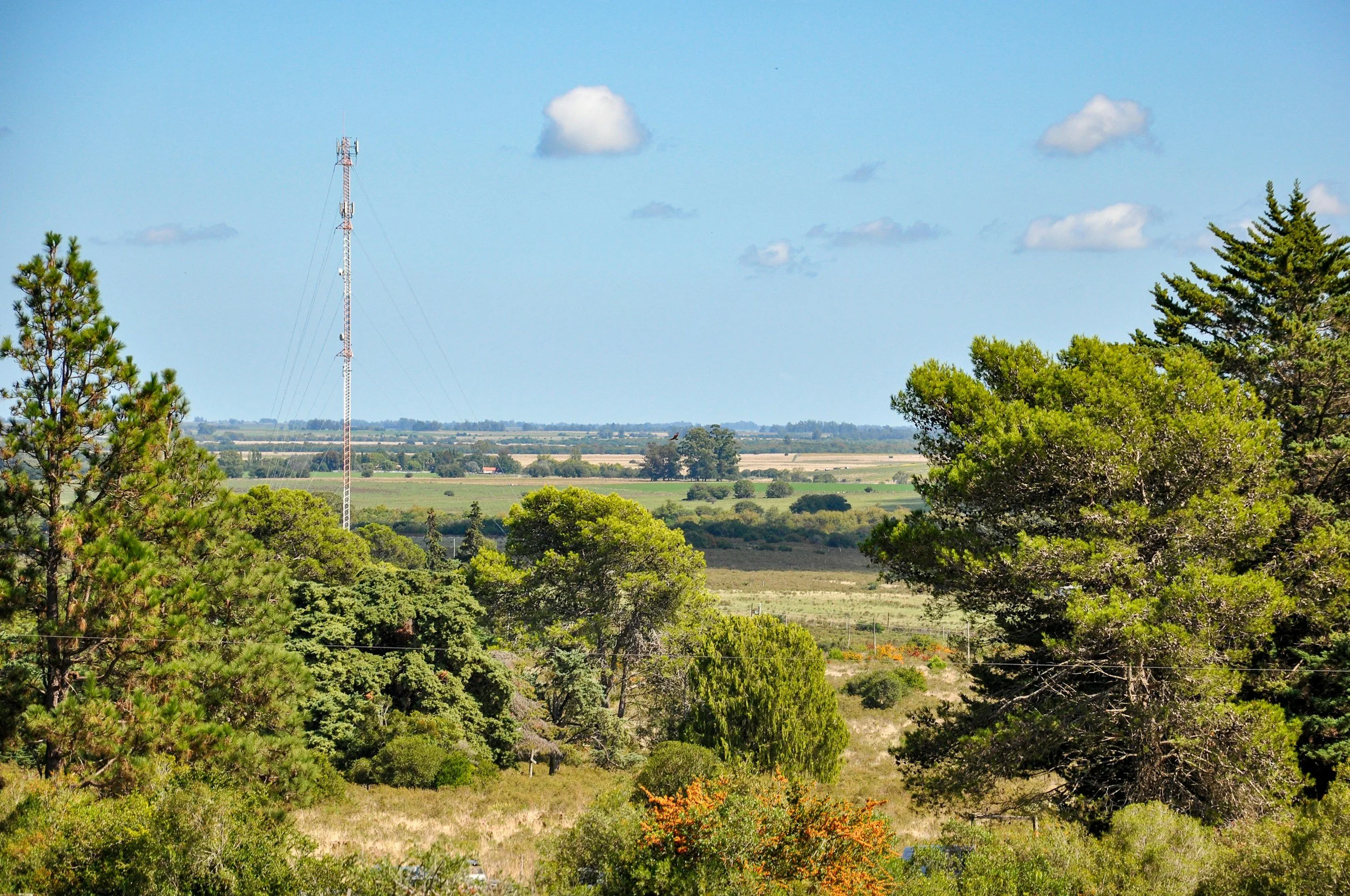 A countryside view with green trees, open fields, a tall communication tower, and a blue sky with scattered clouds.