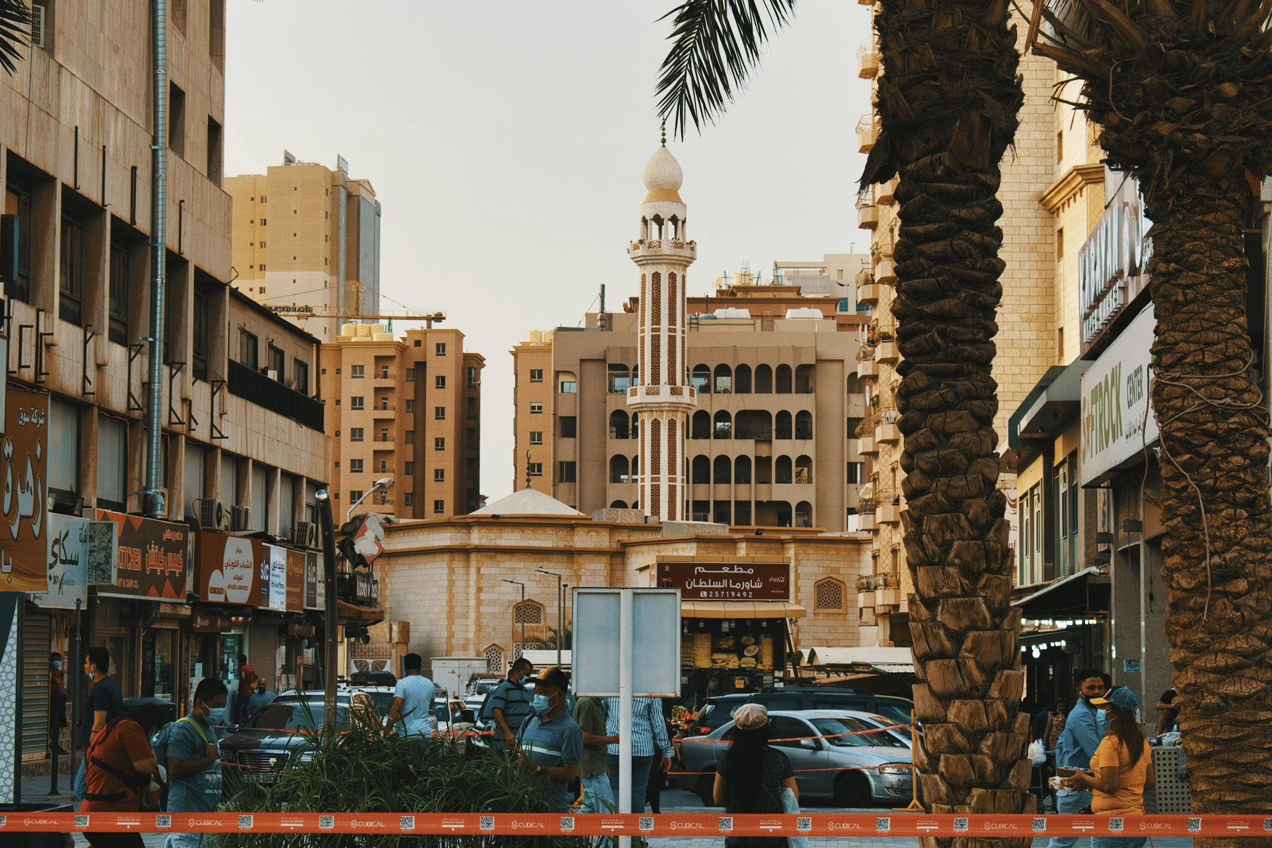 A busy urban street scene with people walking, palm trees, cars, and shops. In the background, there is a mosque with a tall minaret and modern high-rise buildings surrounding it.