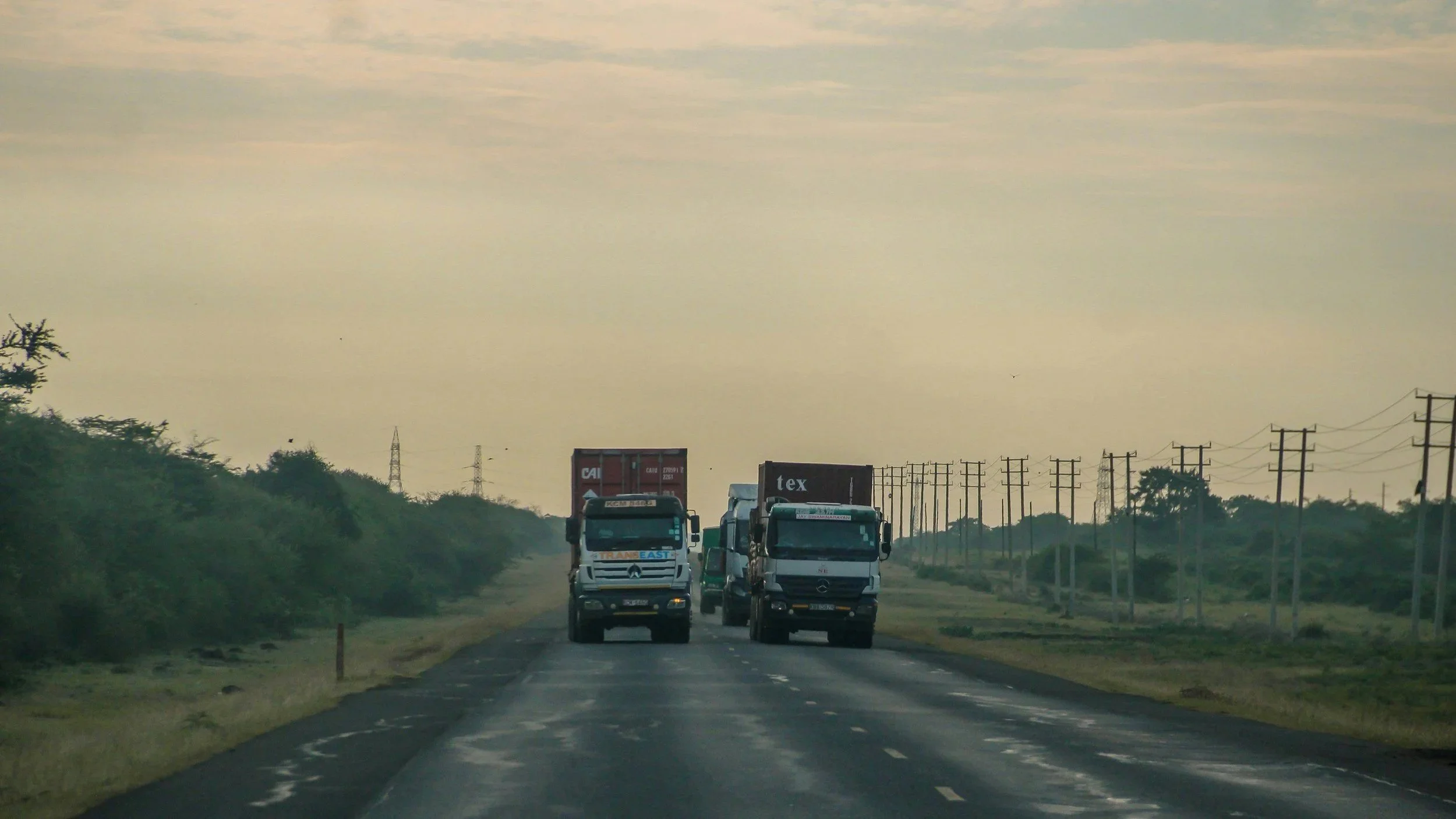 Two trucks driving on a two-lane highway surrounded by green trees and power lines under a cloudy sky.