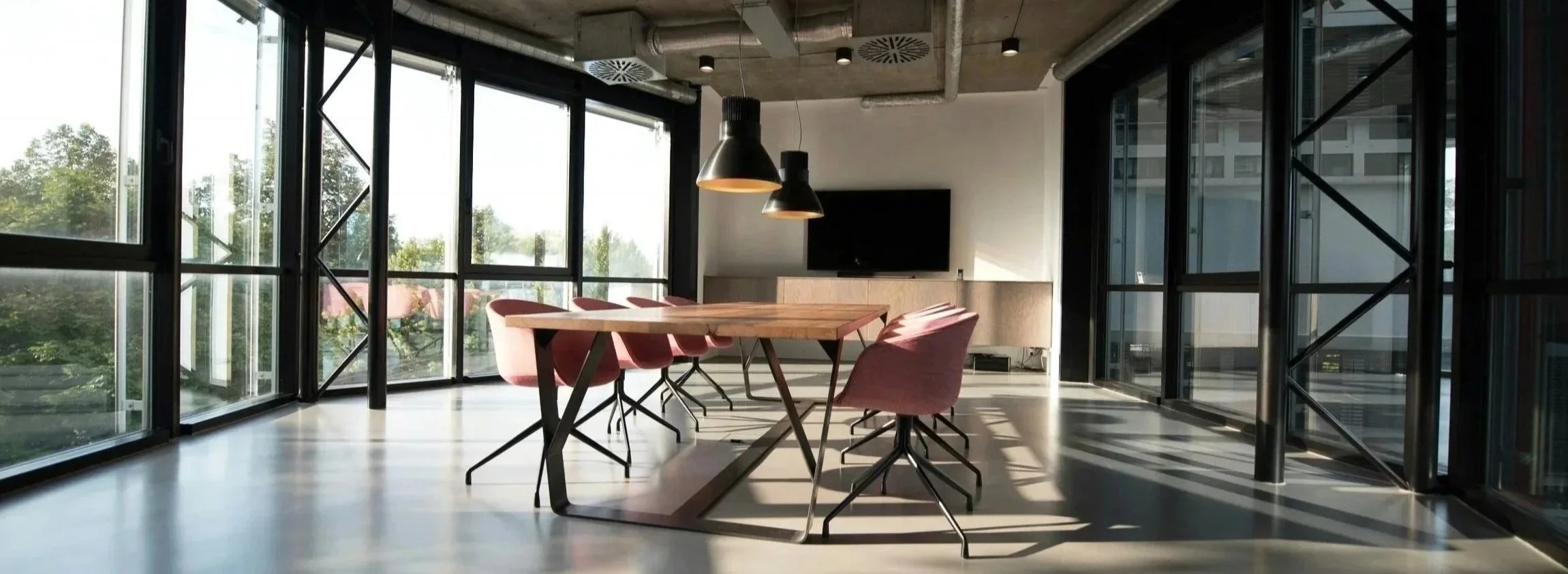 Modern conference room with large glass windows, a wooden table, pink chairs, black pendant lights, and a flat-screen TV on the wall.