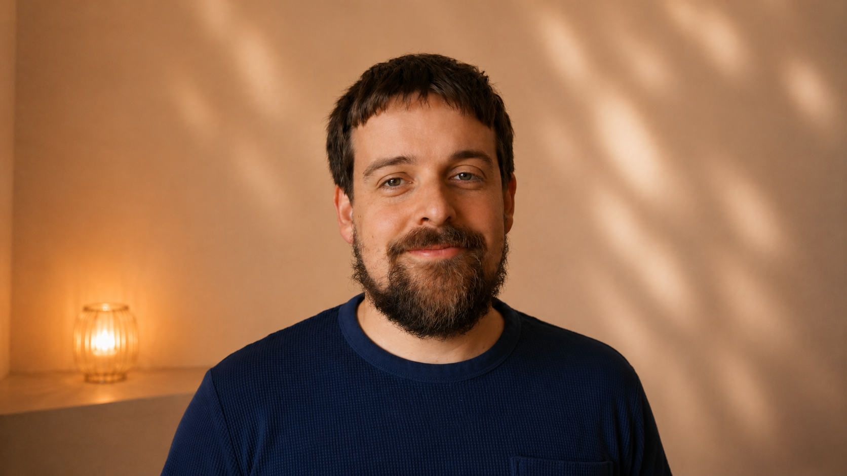 Portrait de François : homme souriant avec barbe, portant un t-shirt noir, debout devant un mur beige avec un éclairage chaud.