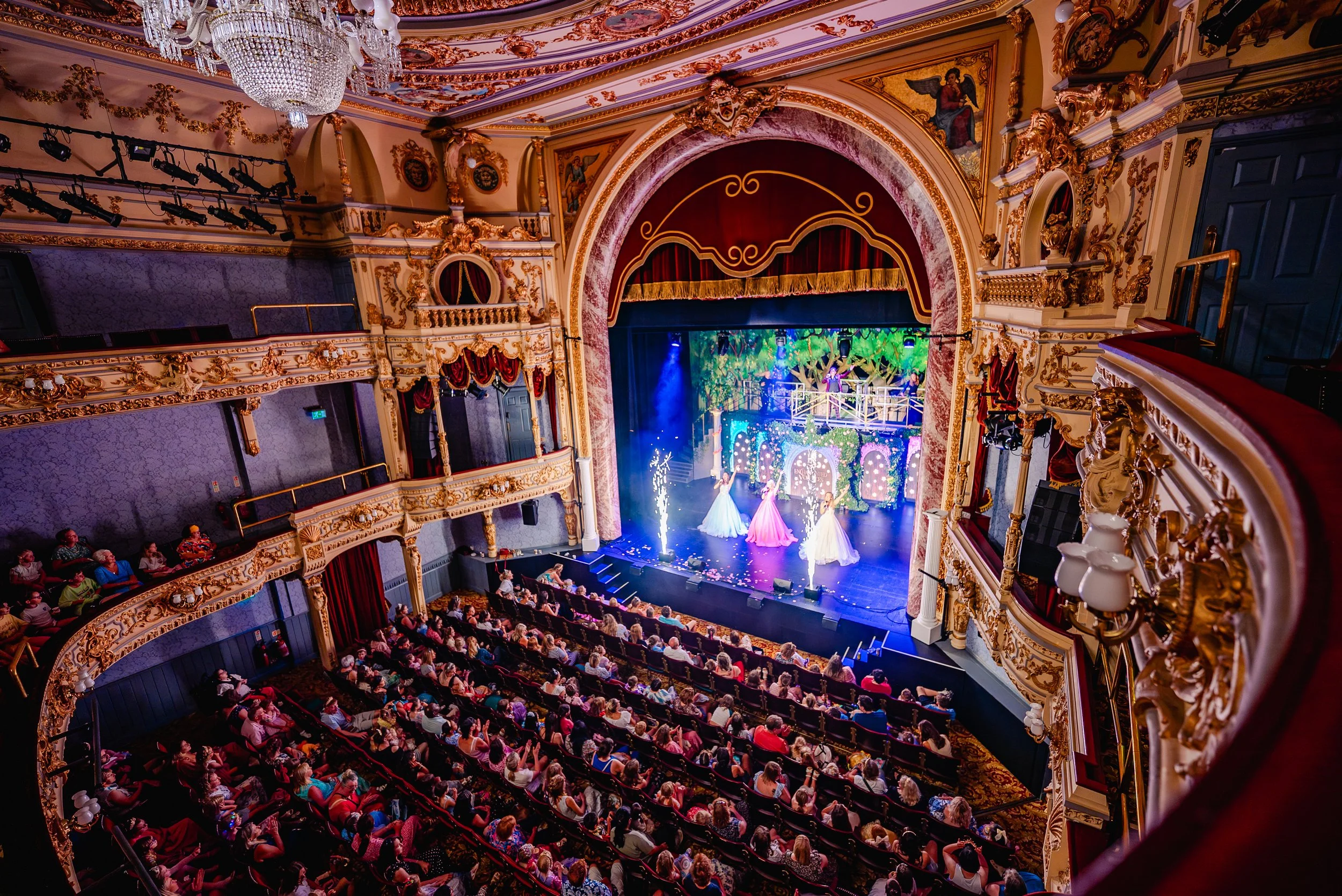 An ornate theater with gold detailing, a large chandelier, and a stage featuring performers in colorful costumes, audience seated in plush seats.