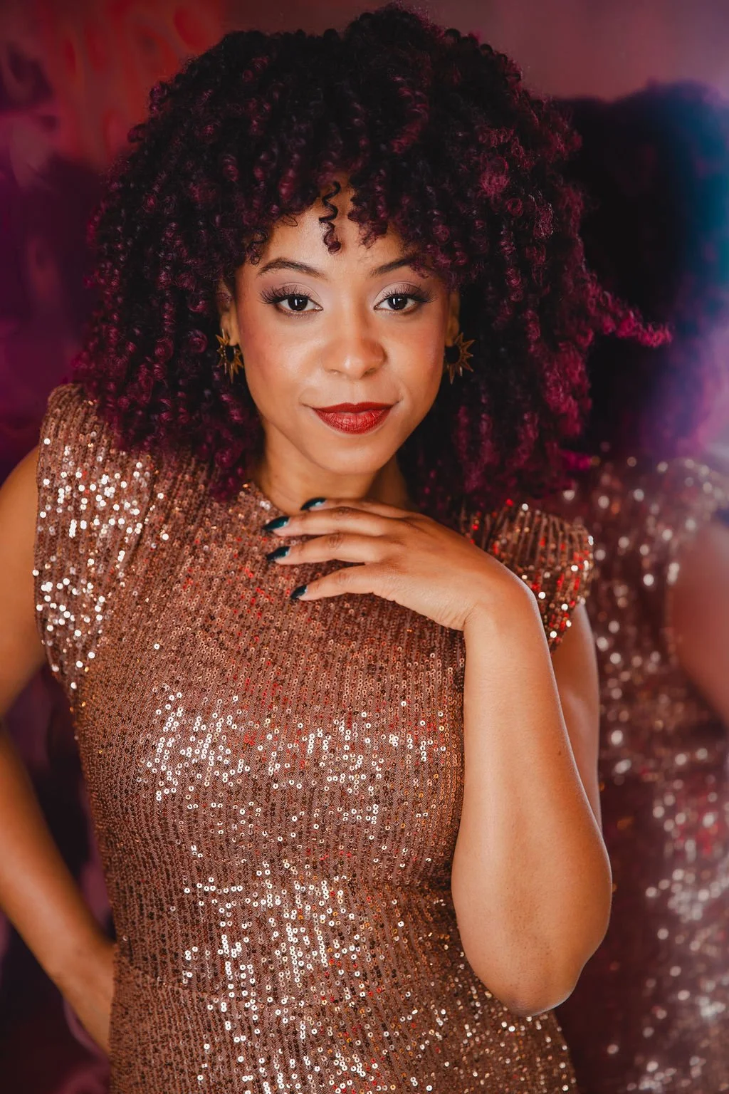 A woman with curly burgundy hair wearing a shimmery, rose gold sequin dress, posing with her hand near her chin and smiling confidently.