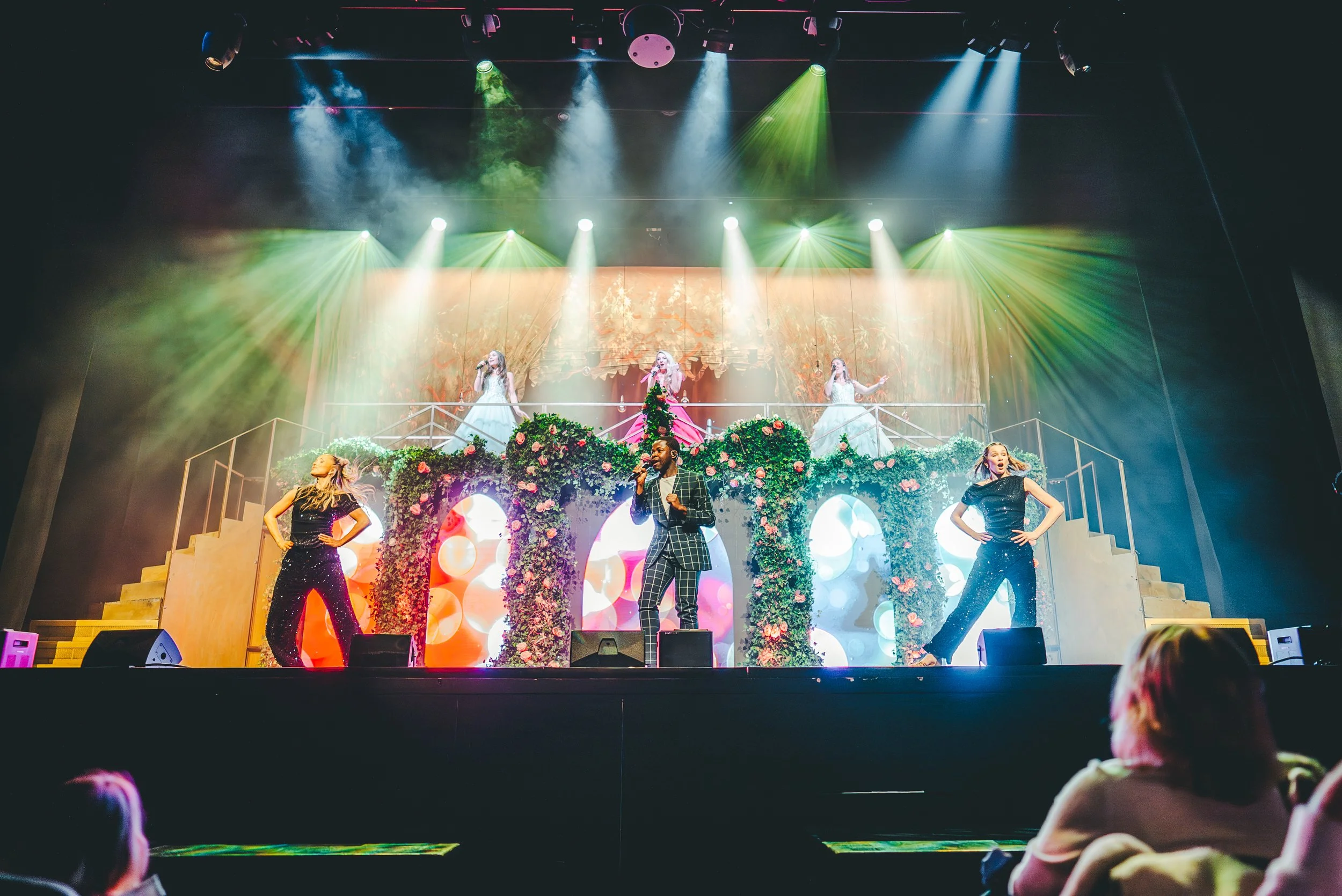 A live stage performance with three women in white gowns singing in front of colorful lighting, and three backup dancers in black outfits dancing in front of a floral and heart-themed backdrop.