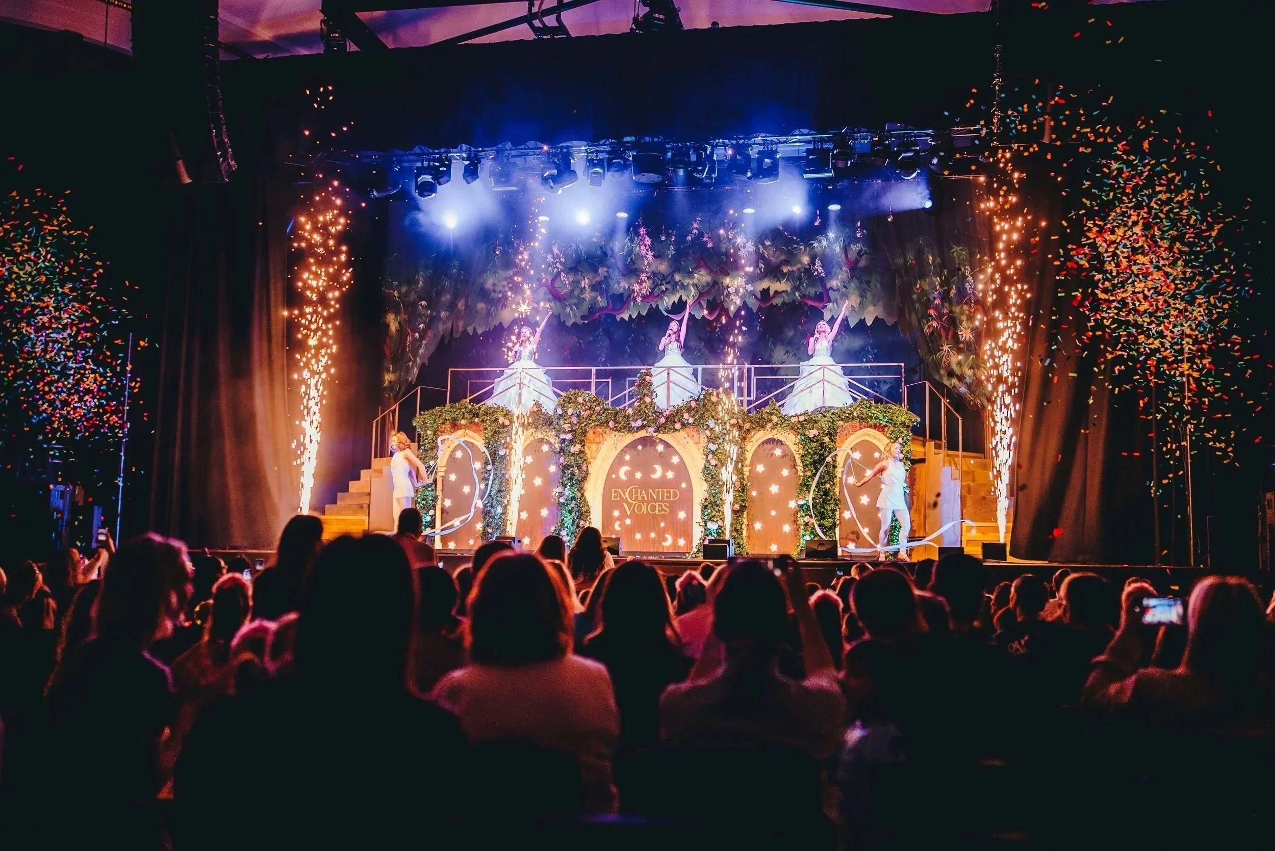 Stage scene from a theatrical performance titled 'Enchanted Voices' featuring three singers dressed in white standing on a decorated platform with flowers and greenery, with fireworks and colorful confetti in the air, and an audience watching in the dark.