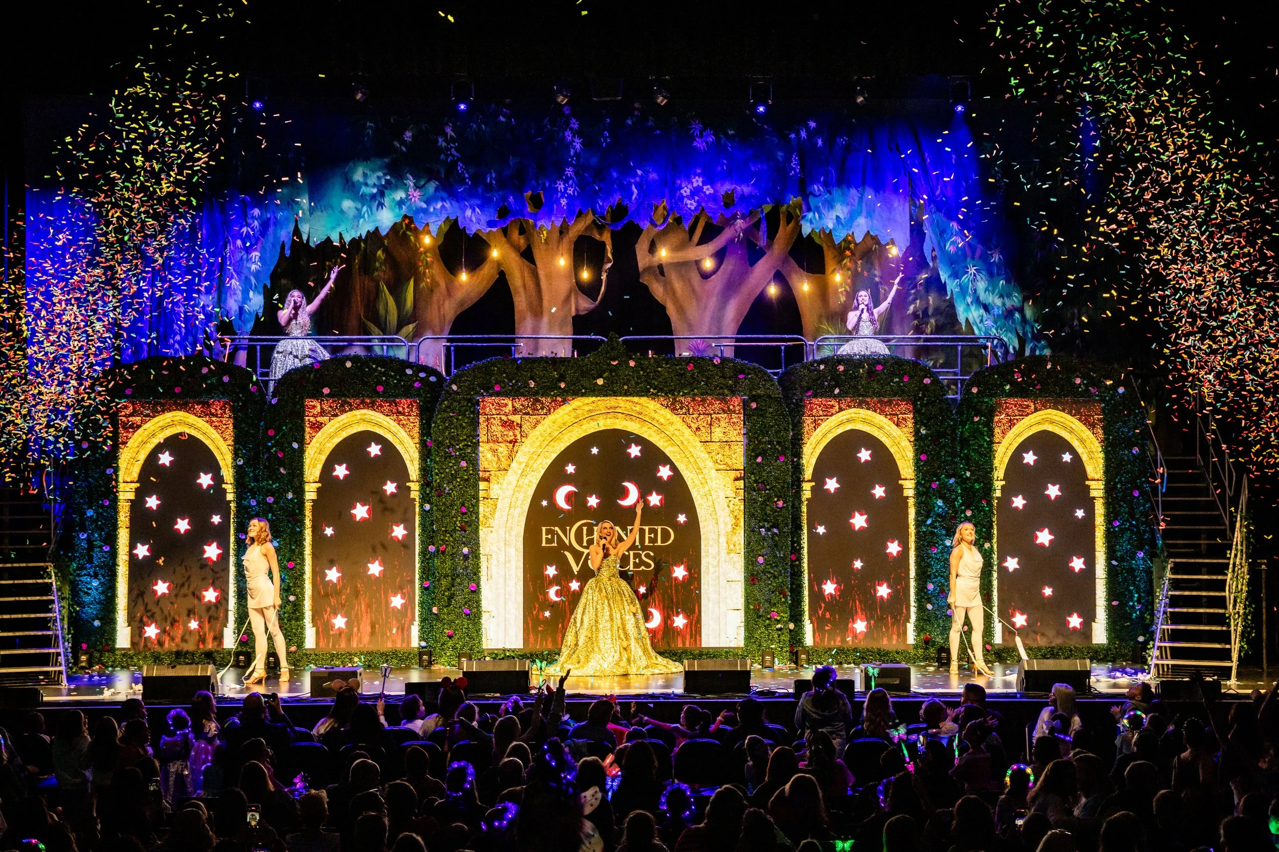 Stage performance with performers in elegant costumes, decorated set with arches, stars, and moons, and confetti falling from above, with an audience watching.